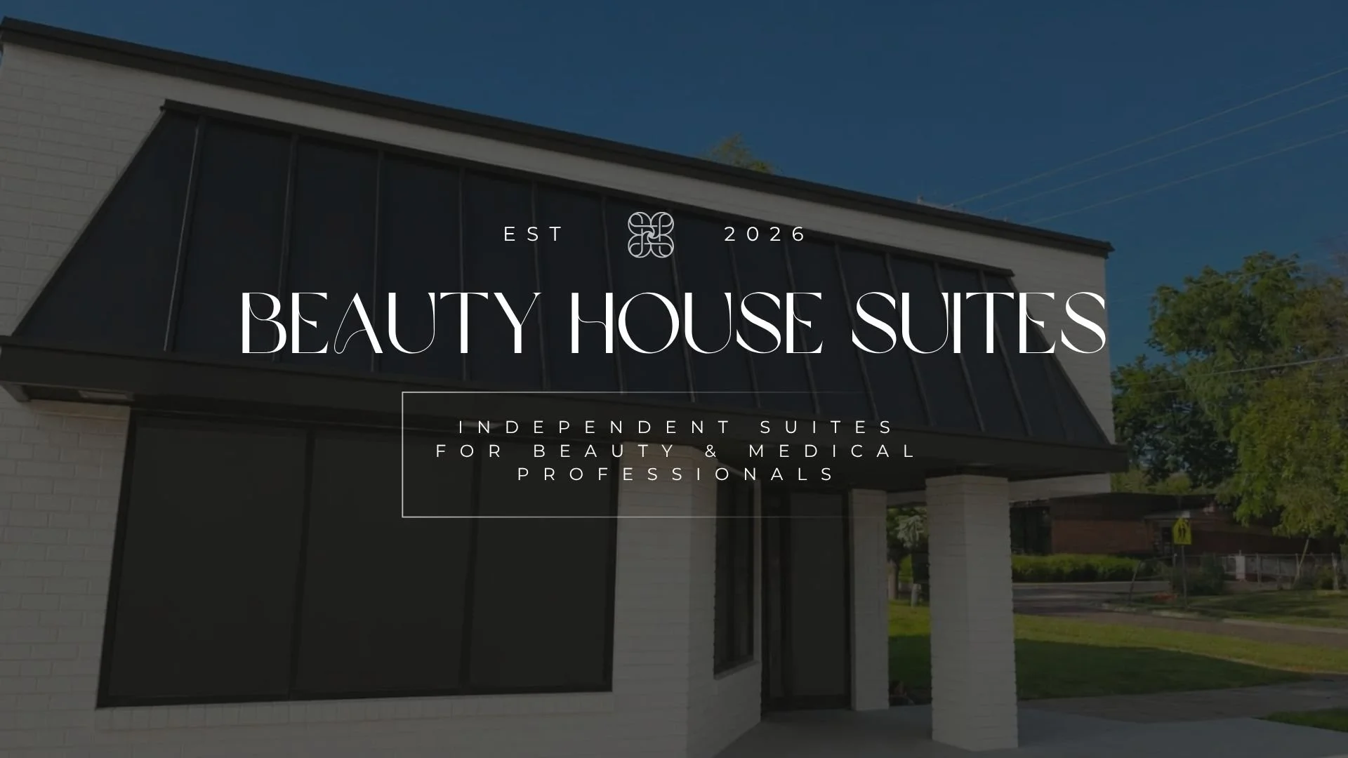 Front of a modern building with black decorative panels and large windows, with trees and a clear blue sky in the background, promoting Beauty House Suites established in 2026, offering independent suites for beauty and medical professionals.