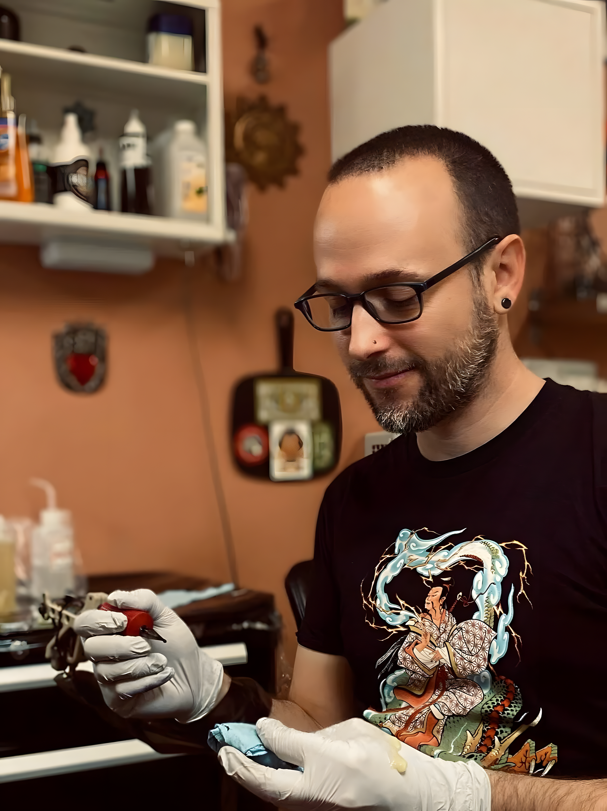 A man with glasses and a beard, wearing a colorful graphic T-shirt, is holding a tattoo machine with gloved hands, working on a tattoo. He is in a room with shelves containing various bottles and supplies, and there are decorations on the wall.