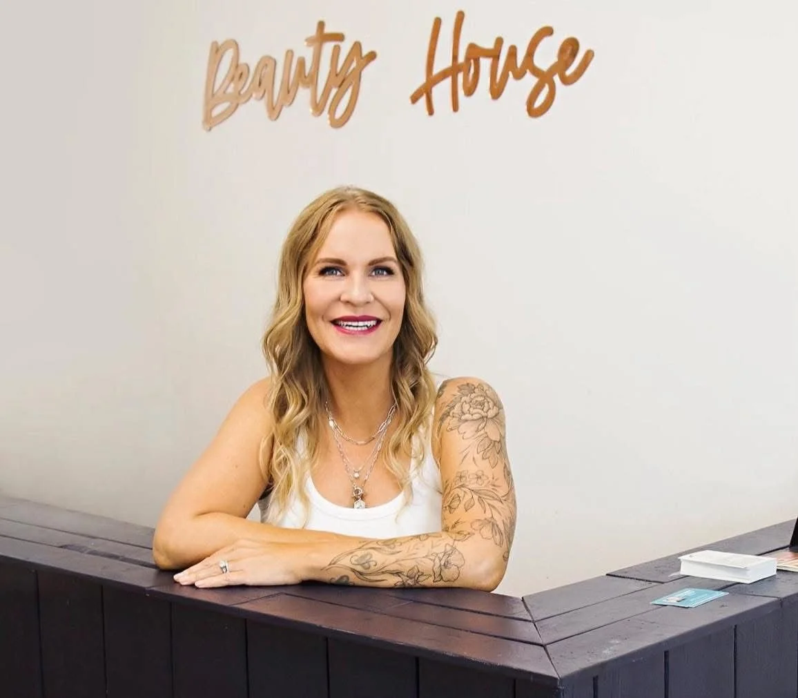 A smiling woman with blonde wavy hair and tattoos on her arms, sitting at a reception desk with a sign behind her that reads 'Beauty House' in script font.
