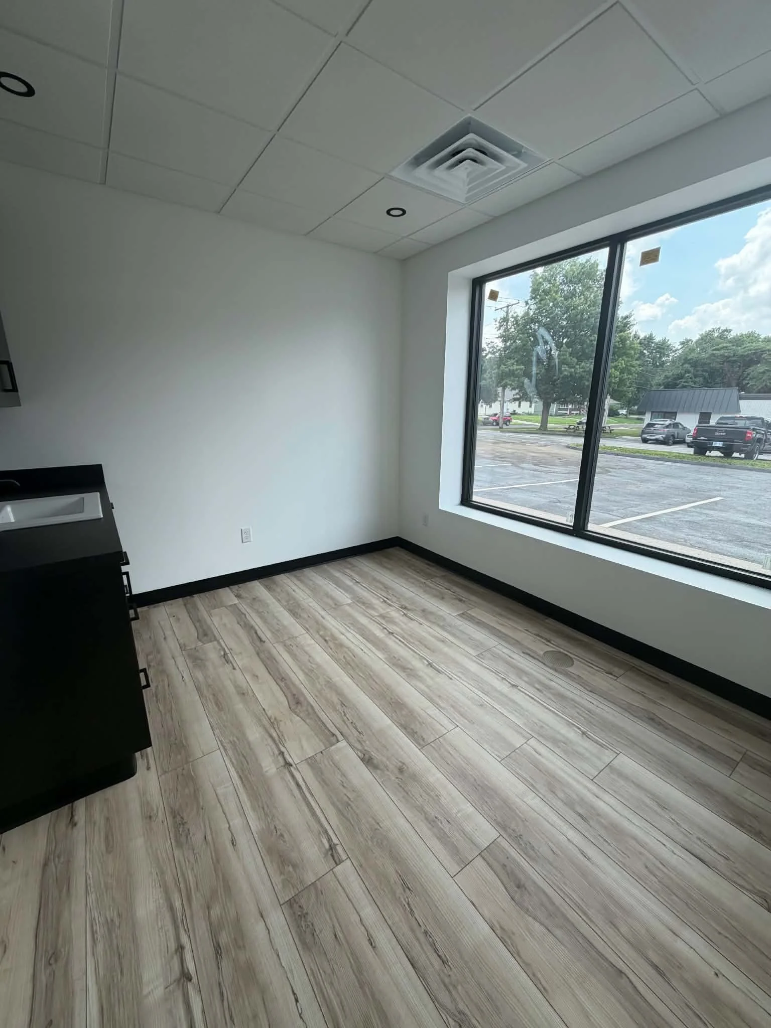 Empty room with wood-patterned flooring, white walls, large window showing parking lot and trees, ceiling with air vent and small recessed lights.