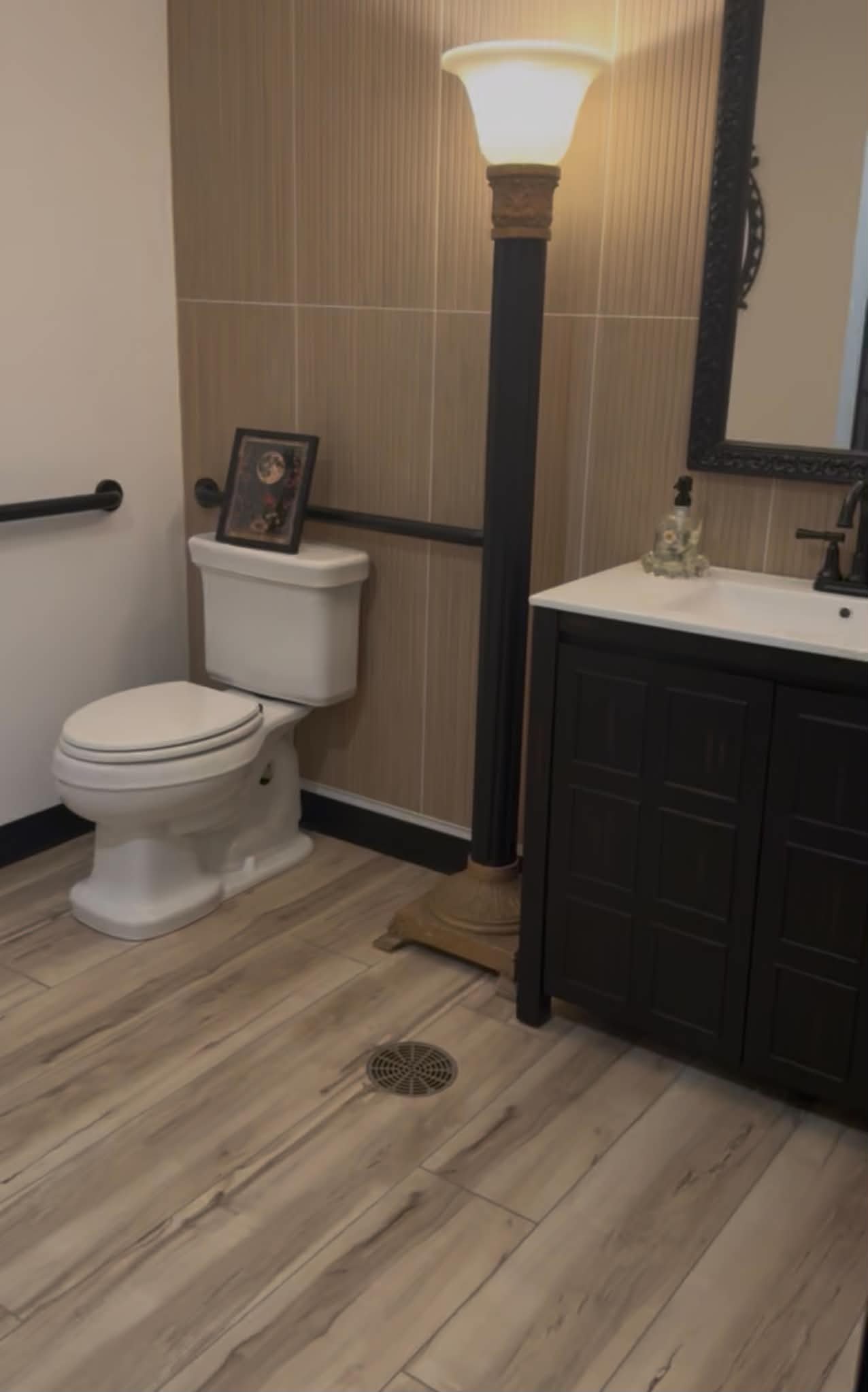 A bathroom with wooden-style flooring, a white toilet with a framed picture on top, a dark wooden vanity with a sink and faucet, a large mirror, a black standing lamp, a hand soap dispenser, a black metal mirror frame, a black grab bar, and a drain on the floor.