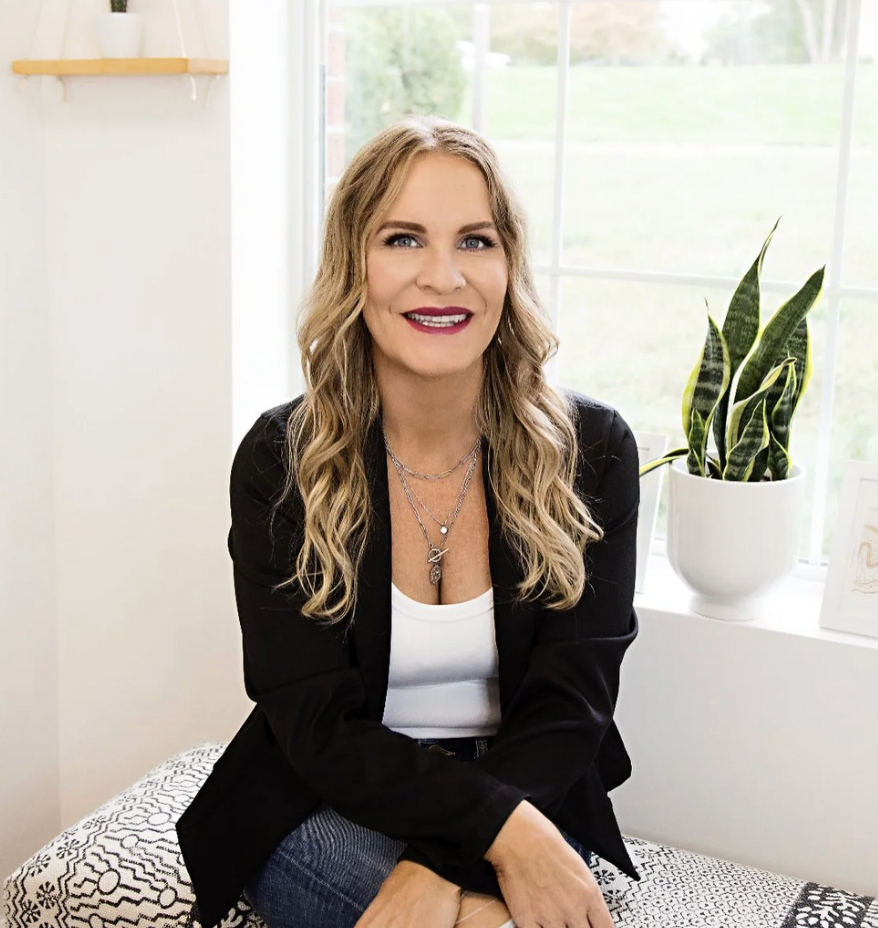 A woman with long blonde hair, wearing a black blazer and white top, sitting on a patterned cushion in a bright room by a window with a potted snake plant nearby.