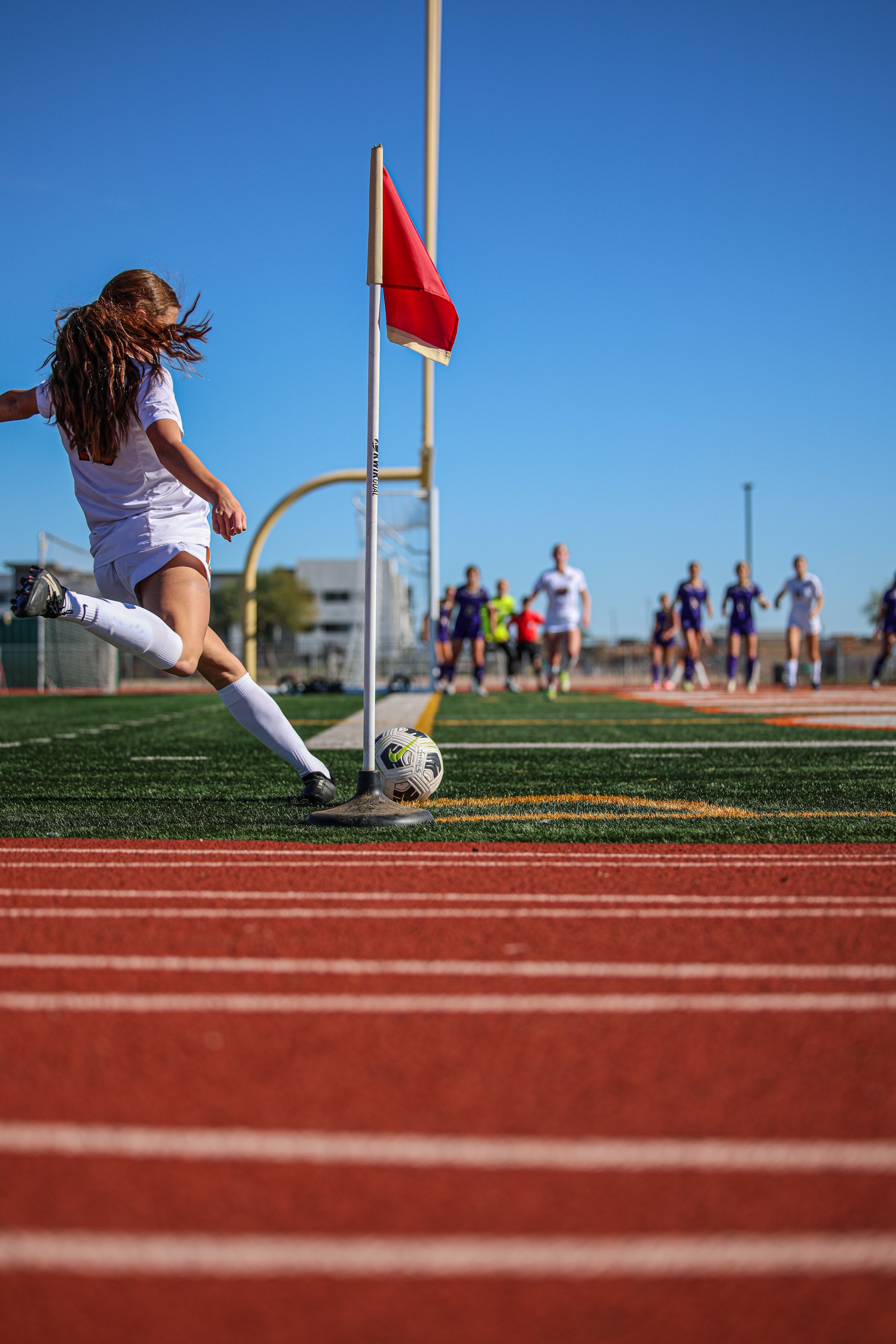 A female soccer player in white uniform kicking a soccer ball near a corner flag on a sports field, with other players in the background under a clear blue sky.