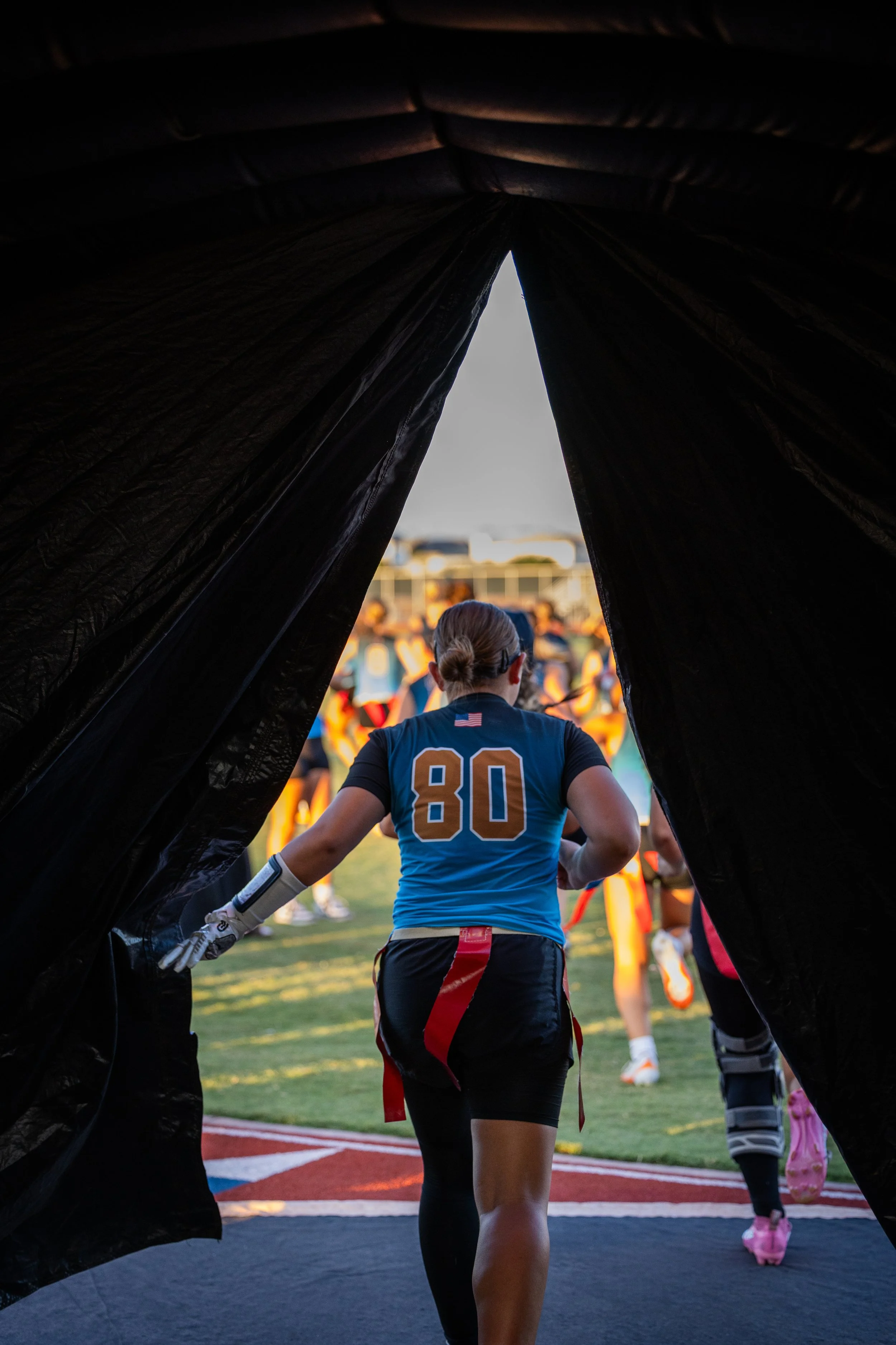 Female football player wearing a blue jersey with the number 80, entering a football field from a tunnel during sunset.