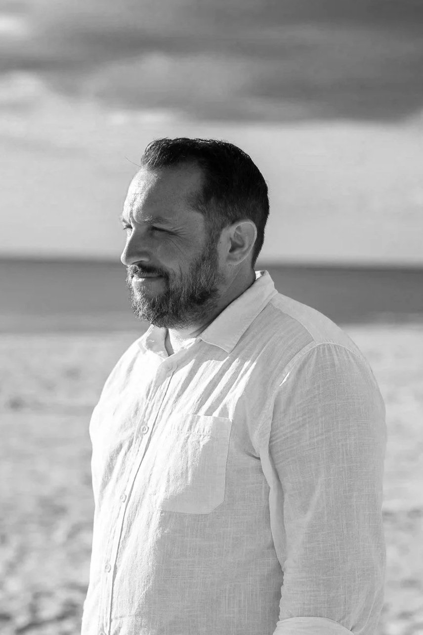 A man with a beard and short hair wearing a light-colored shirt, standing on a beach with the ocean in the background, captured in black and white.
