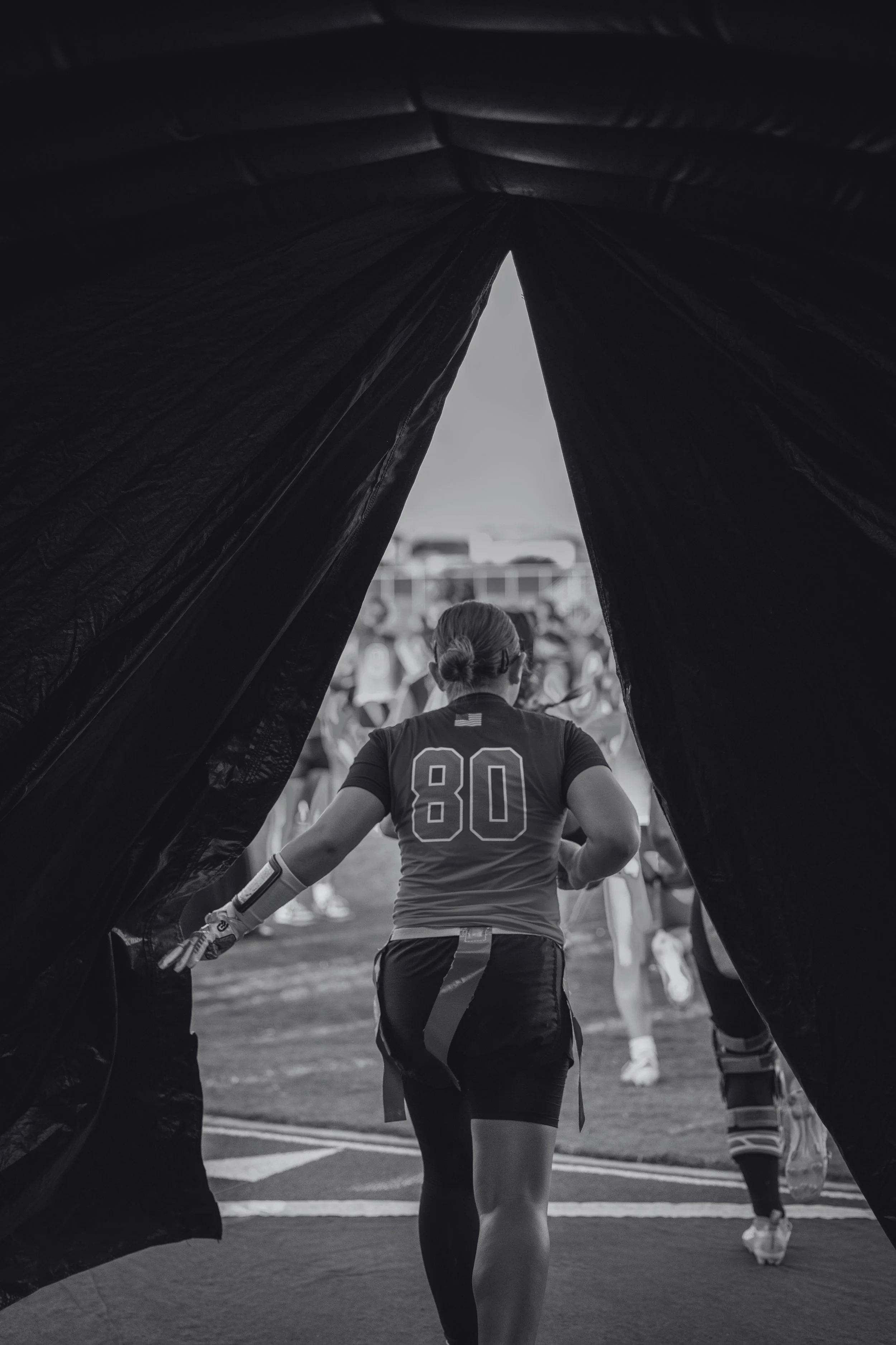 A female athlete wearing a jersey with the number 80 walking onto a sports field through a dark tunnel, with spectators and other players visible in the background.