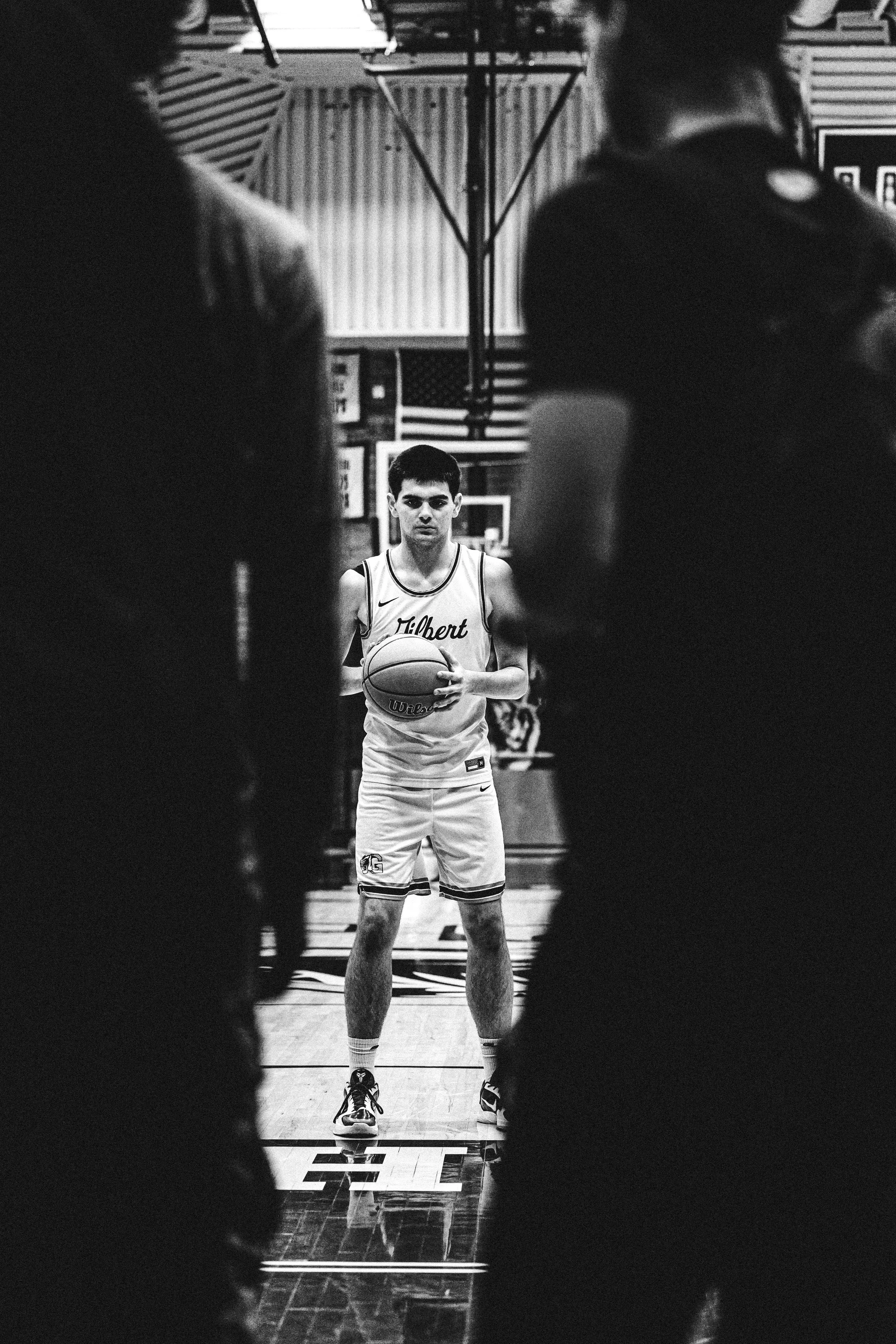 A basketball player stands with a ball at his waist, preparing for a shot during a game or practice in a gym, seen through a gap between two people.