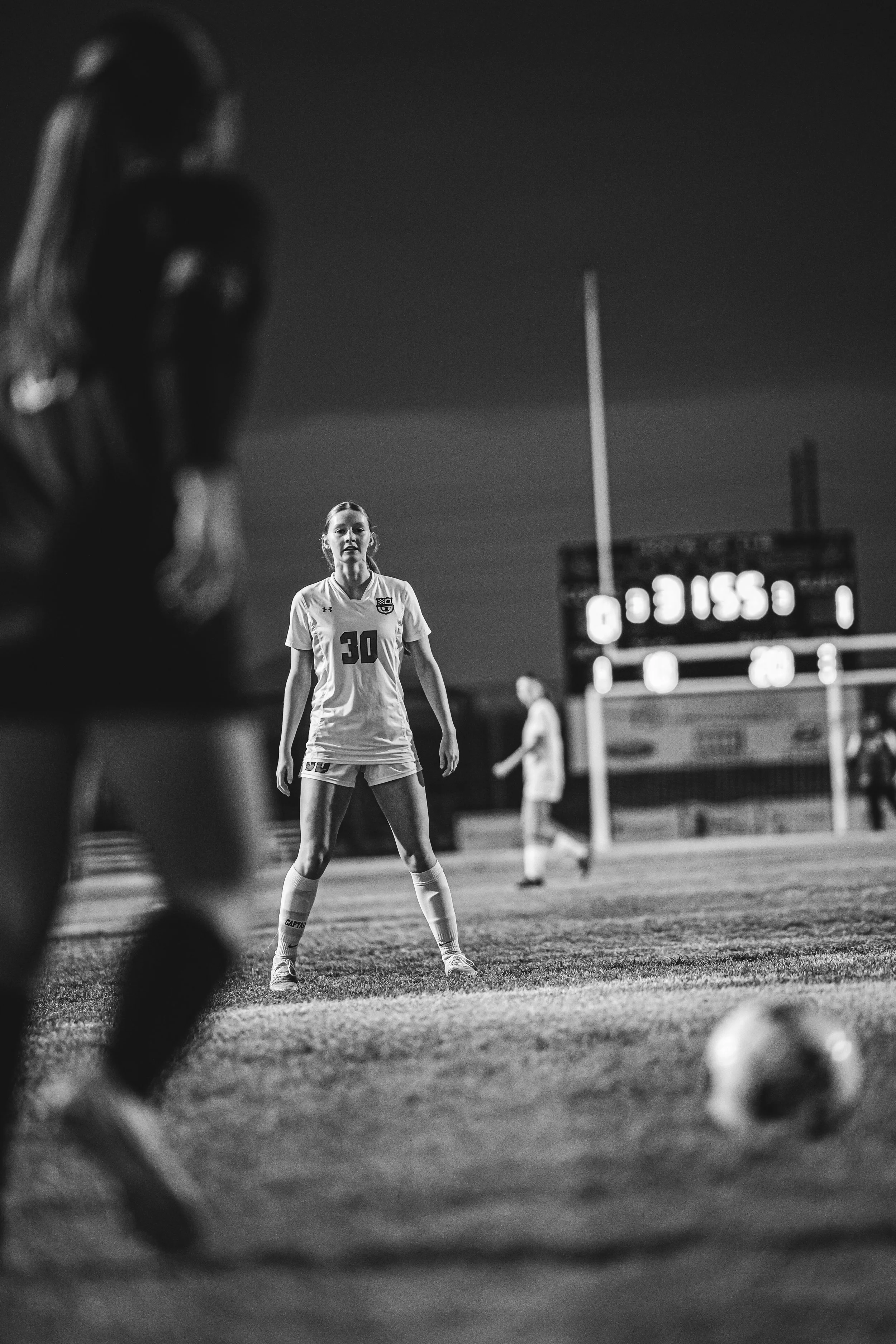 A black and white photo of a female soccer player wearing a jersey with the number 30, standing on a soccer field at night, with a goalkeeper in front of her, a soccer ball on the ground, and a scoreboard in the background.