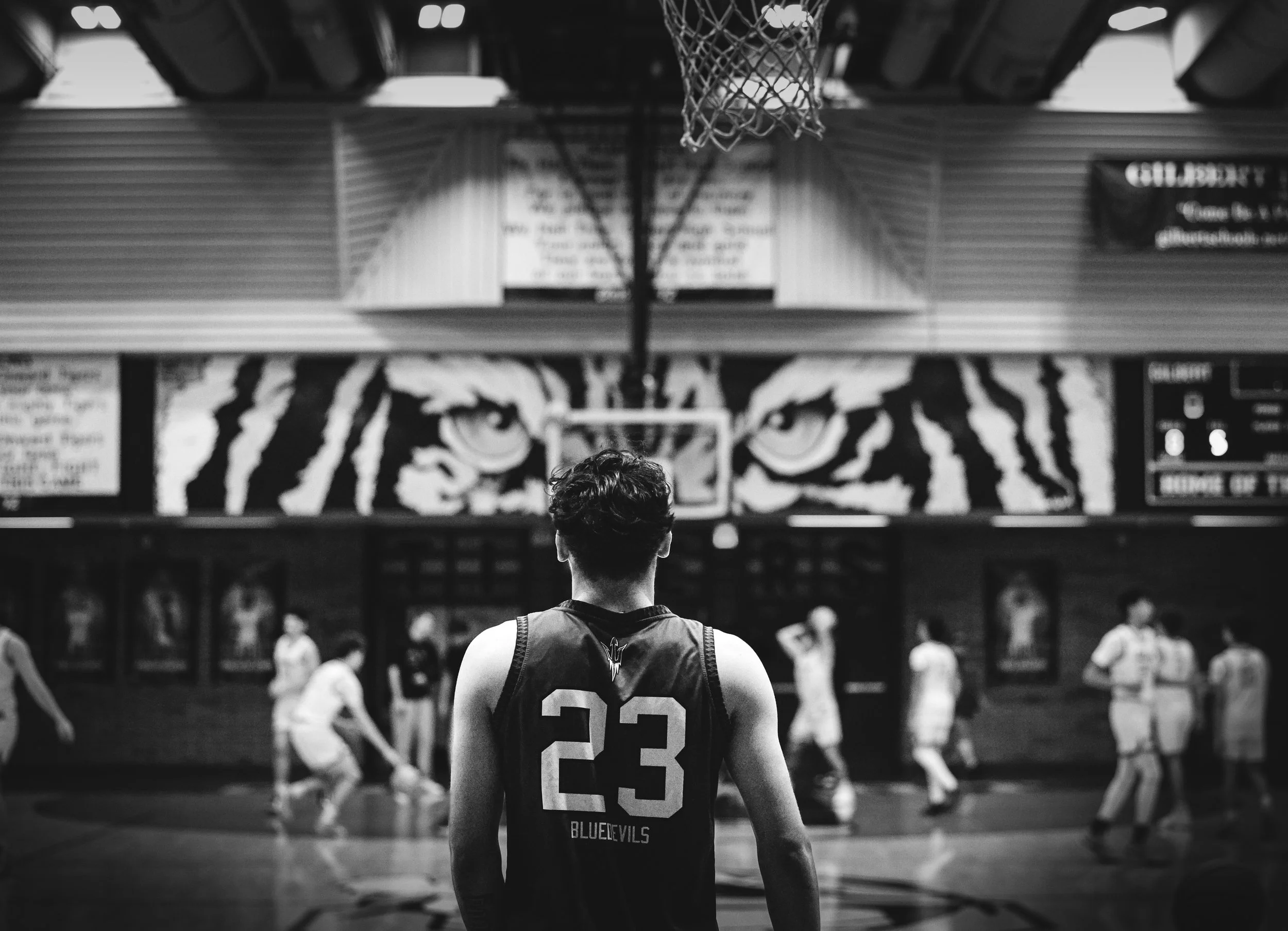 A black-and-white photo of a basketball player with jersey number 23 standing on the court, facing the basket, while other players are in motion in the background inside a gymnasium.