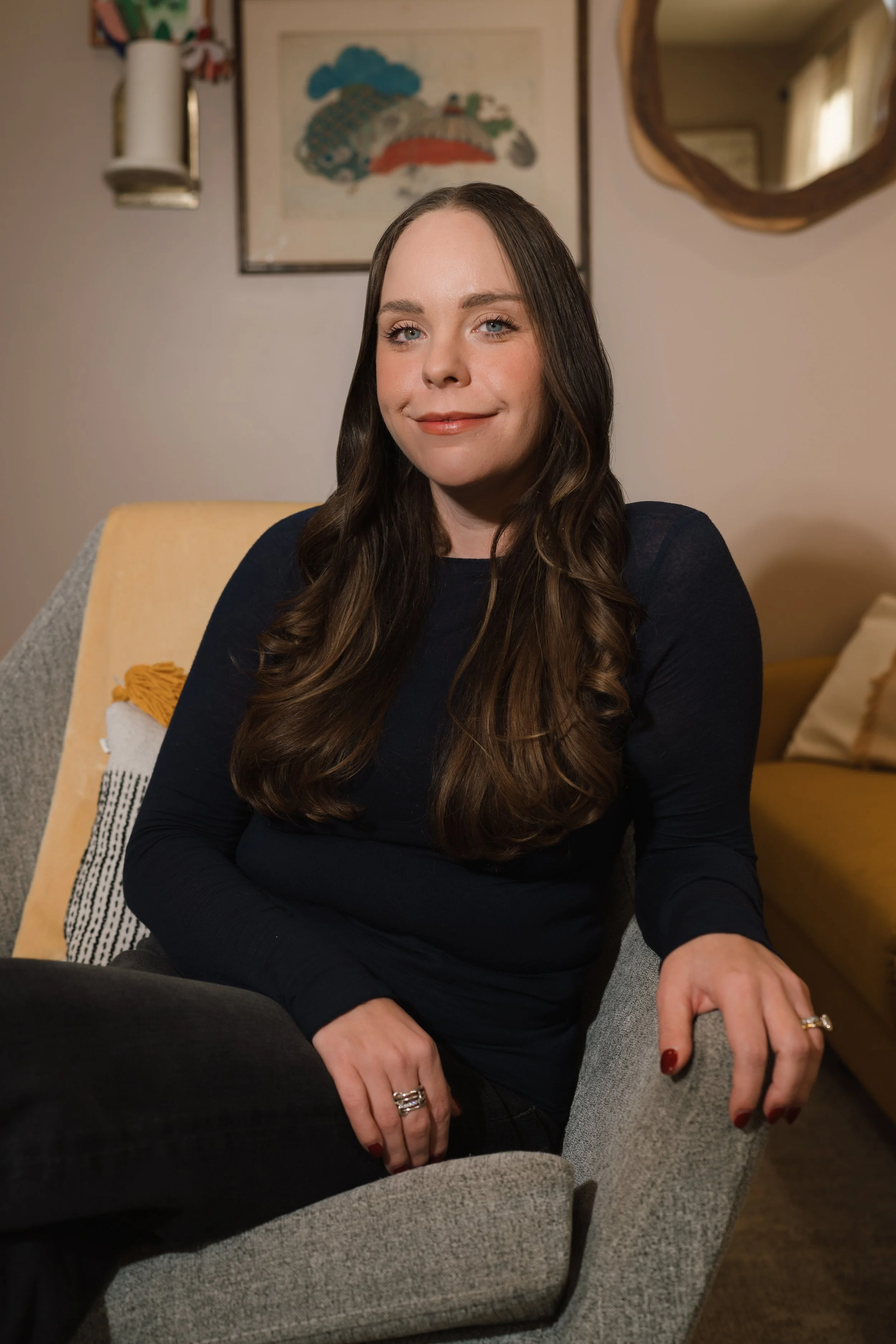A woman sitting in a gray armchair, smiling with long brown wavy hair, wearing a black long-sleeve shirt, in a cozy living room with art and a mirror on the wall behind her.