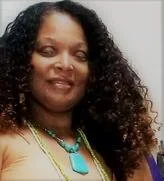 Kenya smiling brightly at the camera. Kenya is a legally blind light-skinned tone Woman of African Descent. She is wearing a nude-colored shirt, a turquoise necklace, and has long blondish-brown hair that is flowing from her right side.   