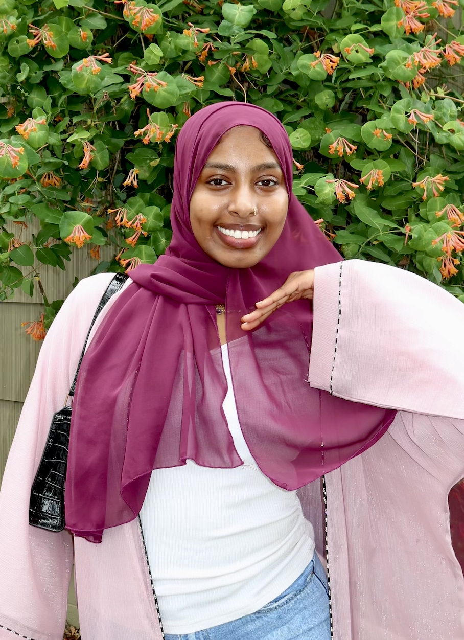 Fateya Omer smiling in a magenta hijab and pink cardigan stands before leafy bushes with orange flowers.
