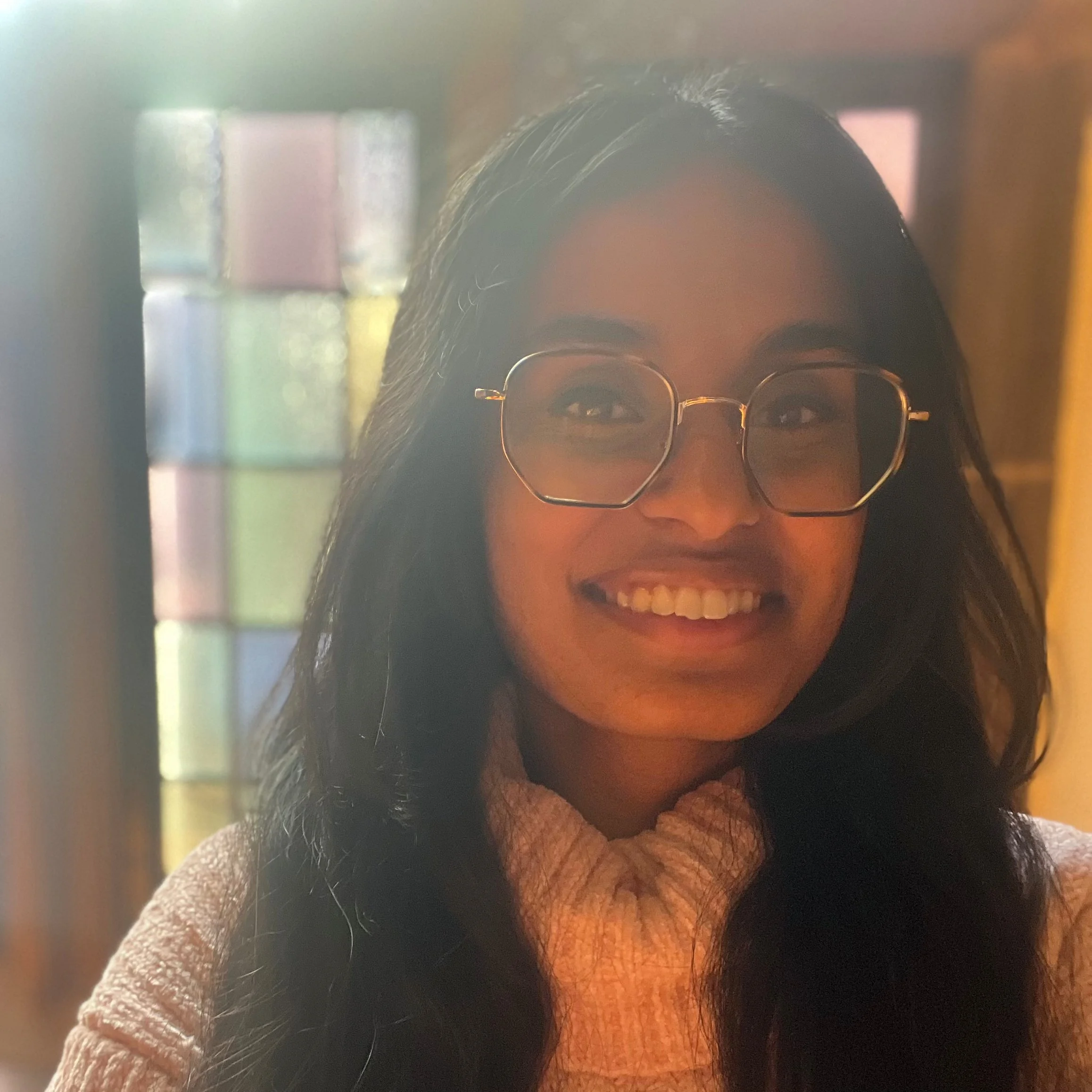 A South Asian American woman wearing large glasses and a cozy pink turtleneck, smiling in front of a stained glass window.