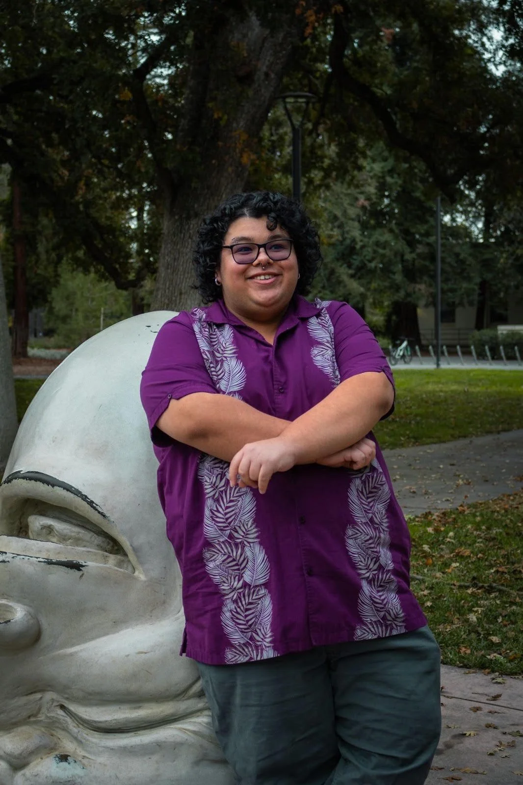 A person with dark, curly hair is facing the camera, smiling. He is wearing gray pants and a short-sleeve purple button-up. He is leaning on a white egg-shaped sculpture in a park-like setting with tall trees, scattered leaves, and a paved walkway. 