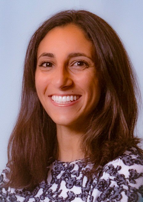 A woman in her early 30s (Kelly) is facing the camera and smiling. She has medium-tan skin, dark brown eyes, and shoulder-length dark brown hair. She is in front of a solid blue background and is wearing a white sweater with navy embroidery. 