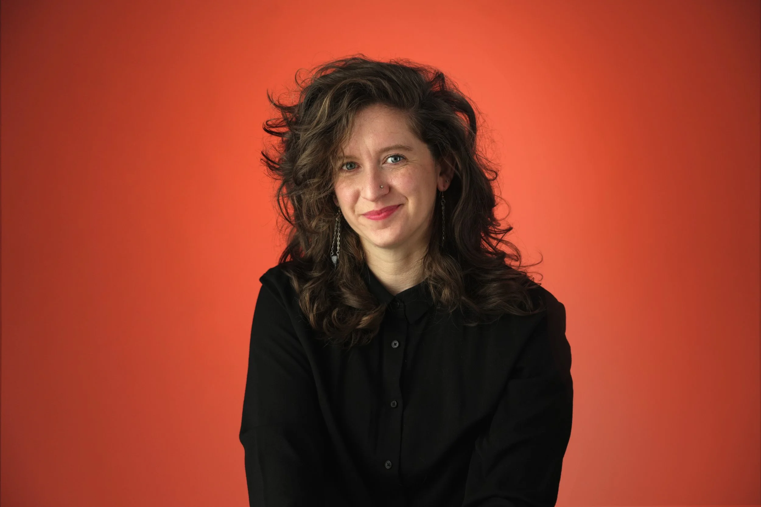 Julia, a white nonbinary femme with wavy brown hair framing their face. They wear a black button-down and dangly earrings and smile cheekily at the camera against a burnt orange background. 