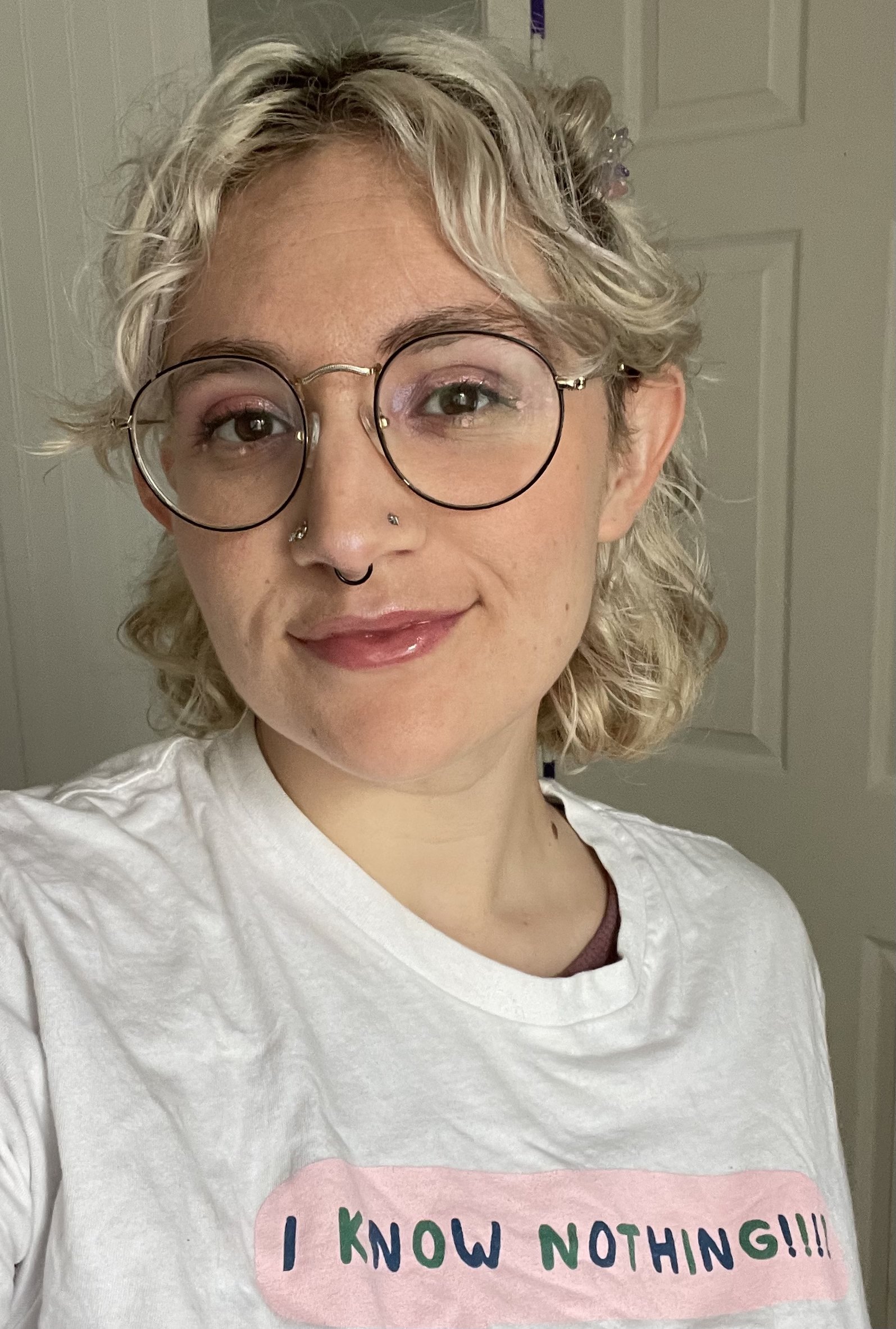 A white nonbinary femme from the chest up looking at the camera. They have platinum blonde hair, black wire-rimmed glasses, two nostril piercings and a septum piercing. They are smiling gently in a t-shirt with the words "I KNOW NOTHING!!!!" printed.
