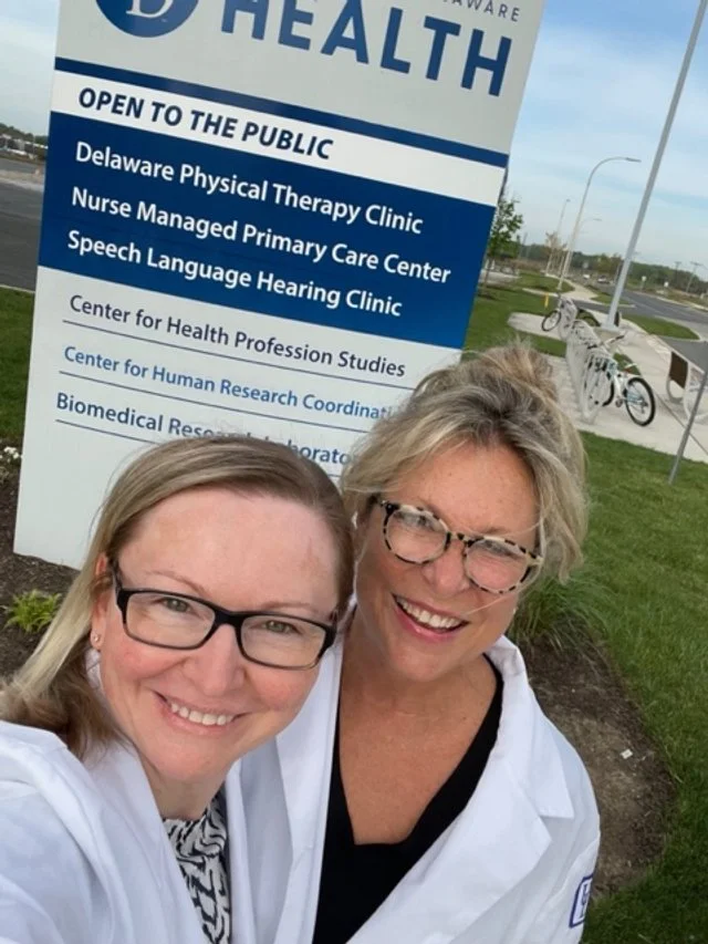 Two women wearing white medical coats taking a selfie in front of a health clinic sign, with bicycles and a road in the background.