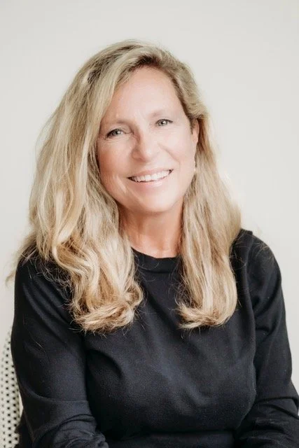 A woman with long, wavy blonde hair smiling, wearing a black top, against a plain light background.