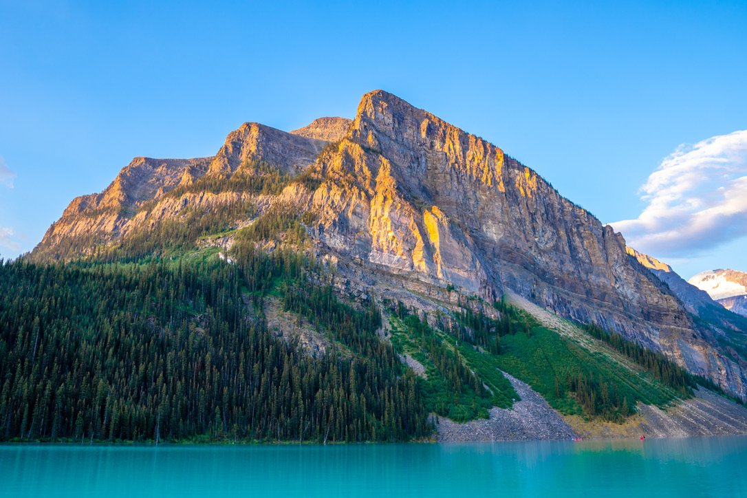 A large mountain next to a body of water, with a forested slope and clear blue sky.