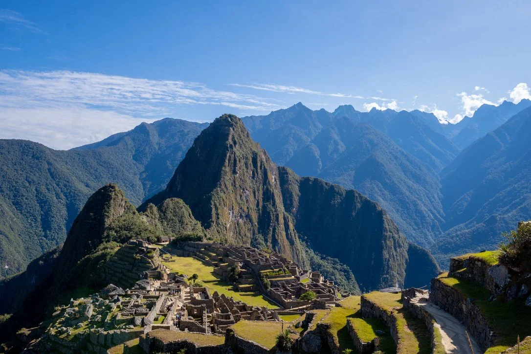 Panoramic view of Machu Picchu, ancient Incan ruins in the Andes mountains with lush green terraces and steep peaks under a blue sky.