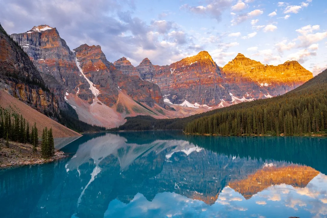 Mountain range with snow patches reflects on a calm blue lake, surrounded by dense pine forest, under a partly cloudy sky at sunset.