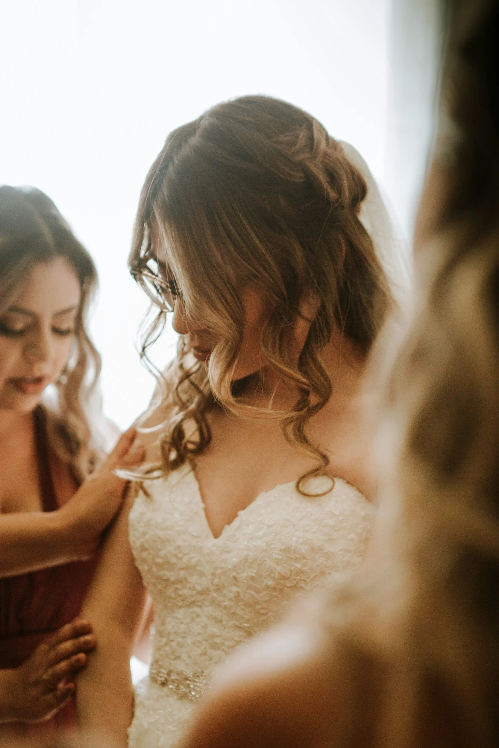 A woman with long wavy brown hair wearing glasses and a white textured wedding dress is being helped by a woman in a reddish dress, possibly preparing for her wedding.