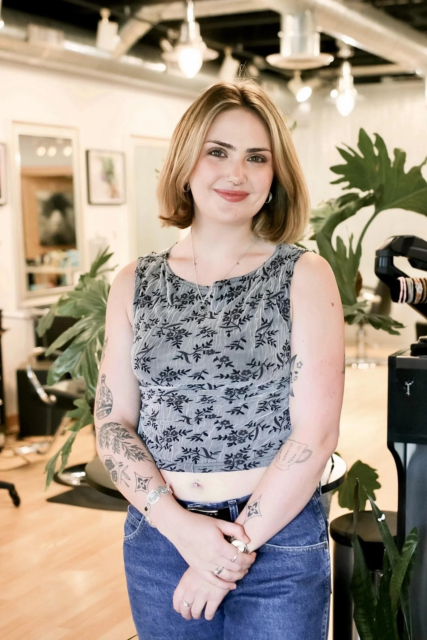 A young woman with shoulder-length blonde hair and a nose ring, wearing a sleeveless gray top with black floral pattern, standing in an indoor room with plants and art on the walls.