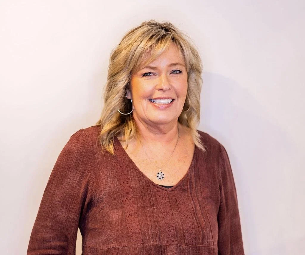 A smiling woman with blond hair, wearing a maroon top and jewelry, standing against a white background.
