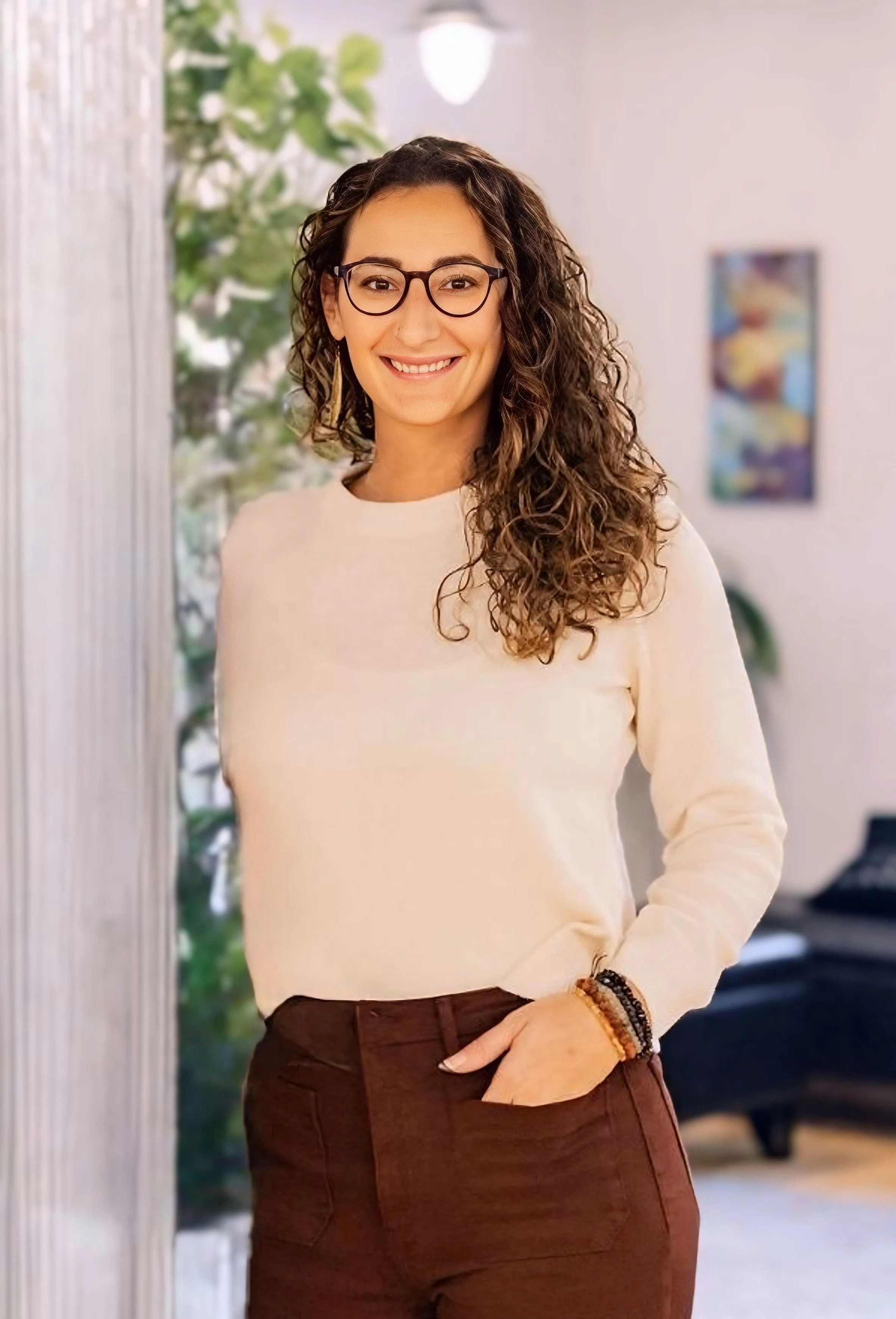 A smiling woman with curly hair glasses and a beige sweater standing indoors near a window and a blurred colorful art piece in the background.