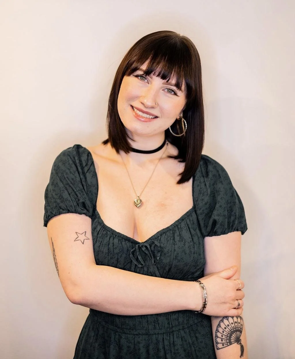 A smiling woman with dark brown hair, wearing a black dress, gold jewelry, and tattoos on her arm, standing against a plain background.
