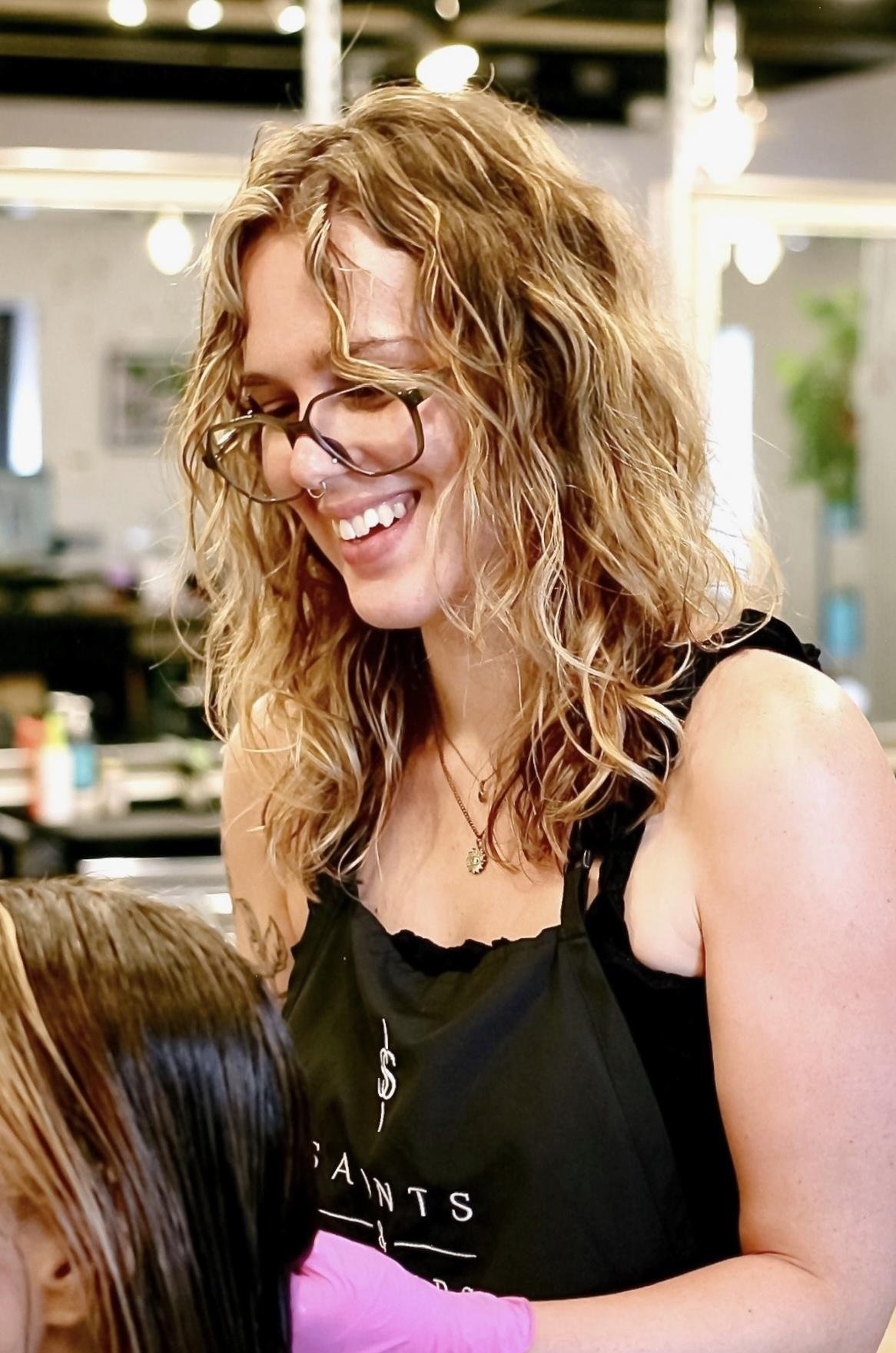 A woman with curly blond hair, glasses, and a nose ring smiling, wearing a black apron, at a hair salon.