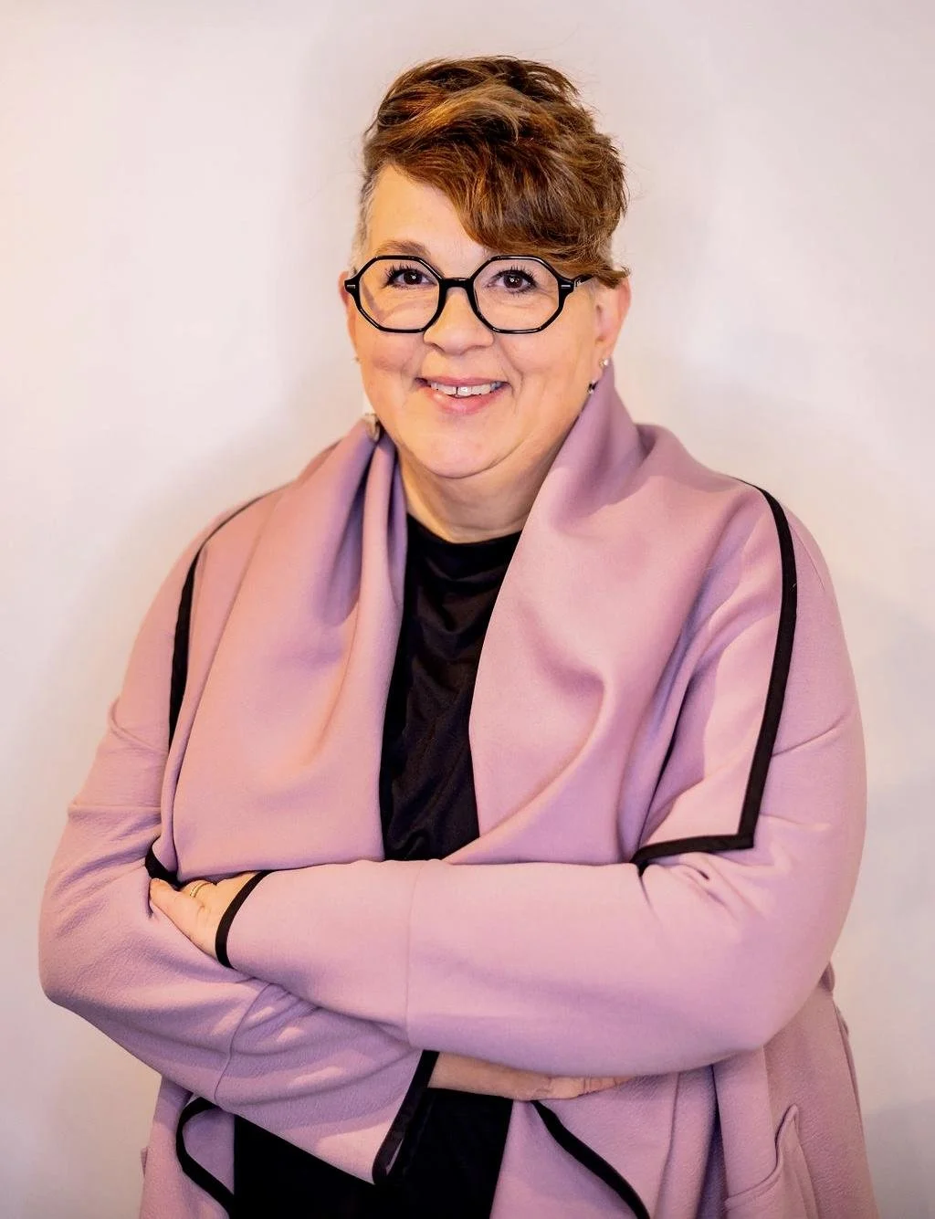 A woman with short, curly hair and glasses, smiling, wearing a black top and a pink blazer with black trim, standing against a plain background.