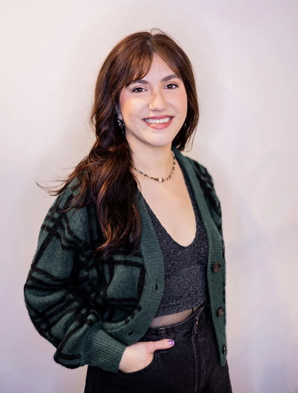 A young woman with long brown hair and light skin, smiling, wearing a green and black plaid cardigan over a black glittery crop top, black high-waisted jeans, necklace, and earrings, standing against a plain light-colored background.