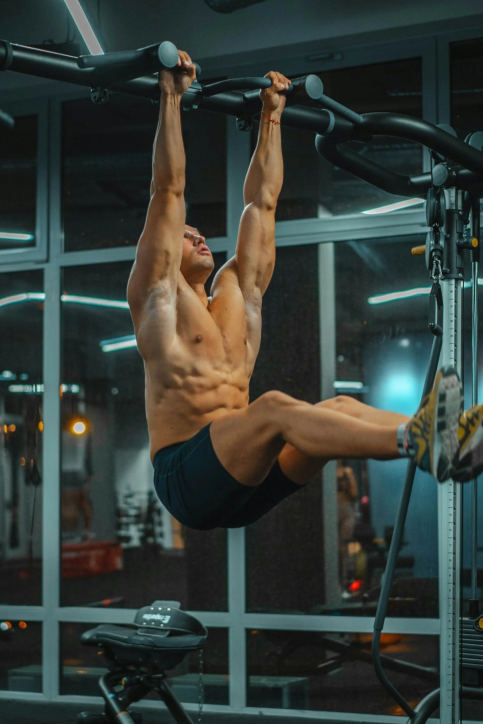 A muscular man performing a hanging leg raise exercise on a pull-up bar in a gym.