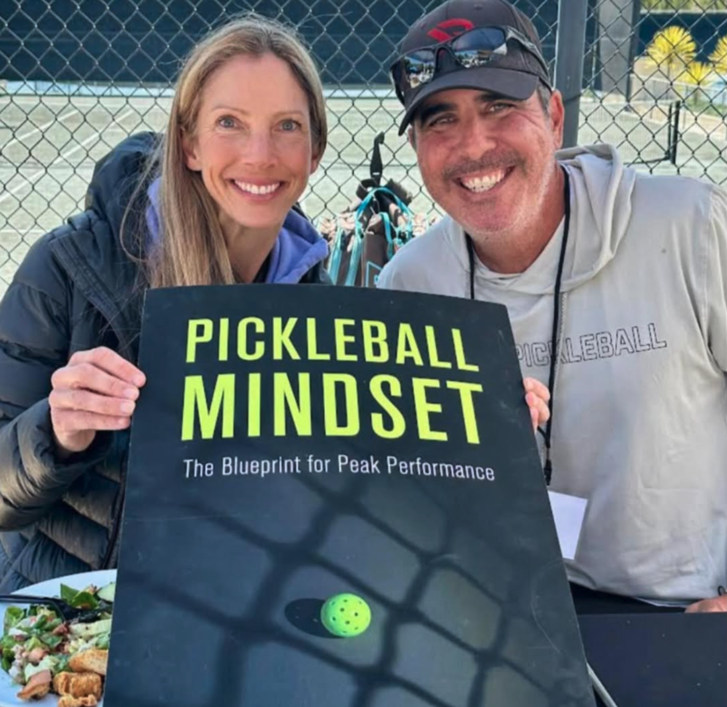A woman and a man smiling in front of a book titled 'PICKLEBALL MINDSET'.