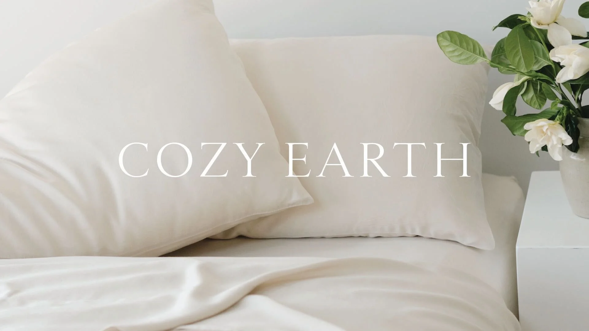 A cozy bed with white pillows, cream-colored sheets, and a vase of white flowers on a nightstand beside the bed.