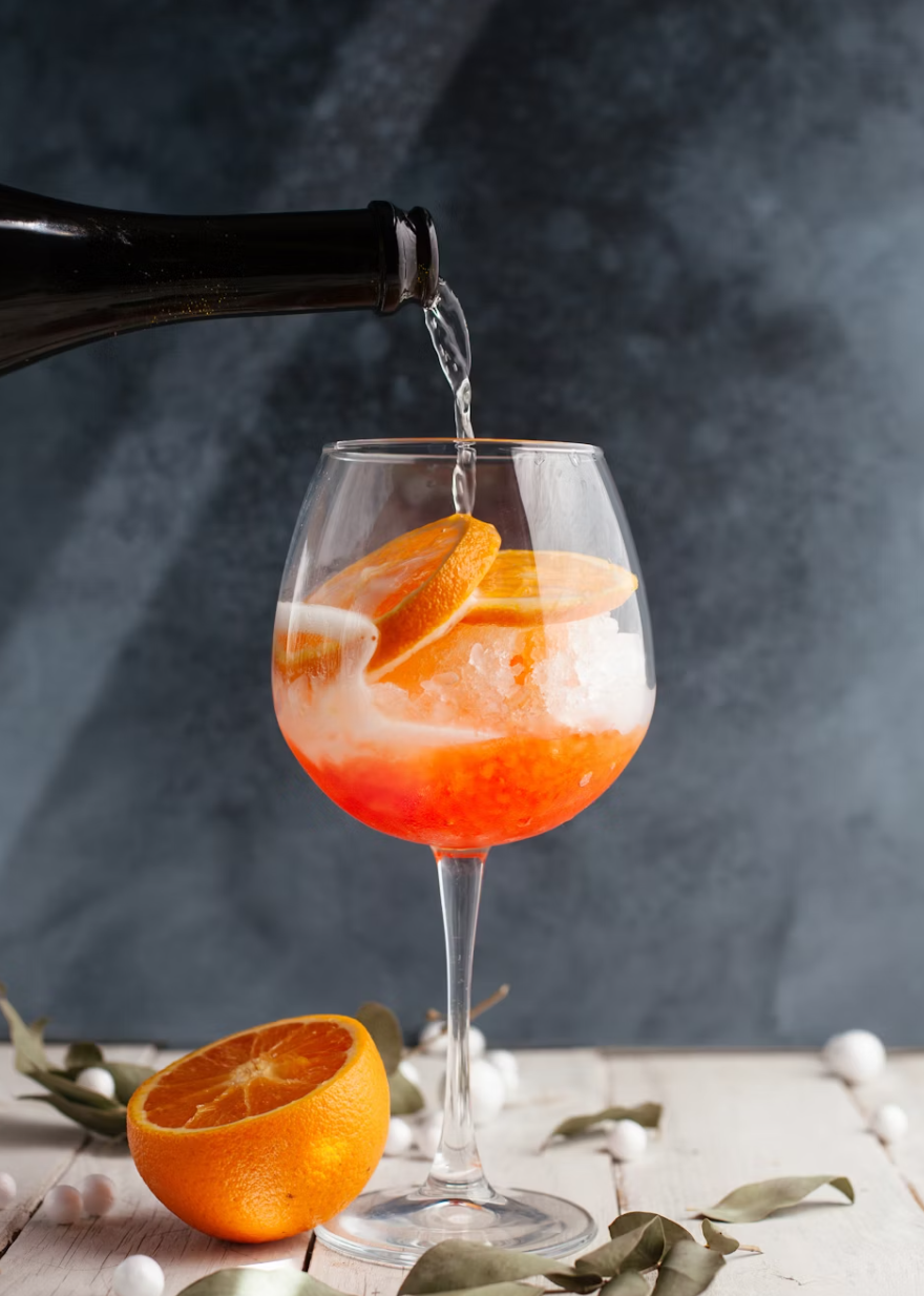 A wine glass filled with a pink-orange cocktail garnished with slices of orange. A bottle is pouring clear liquid into the glass. There is a halved orange and some leaves and white berries on a white wooden surface in front of a dark background.