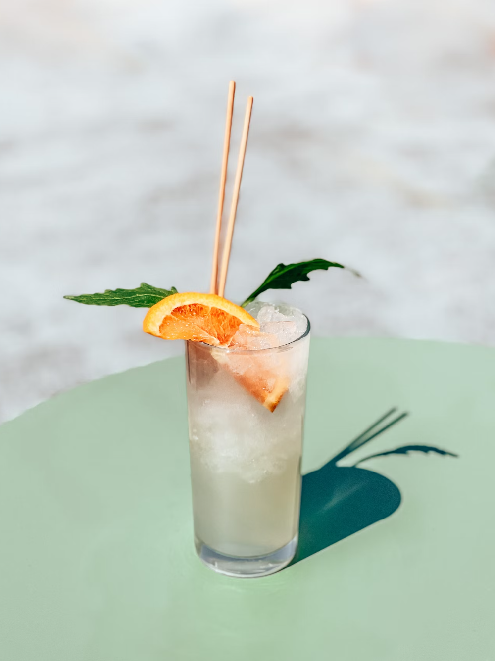 A tall glass of iced cocktail garnished with a slice of orange, two green leaves, and two wooden cocktail stirrers, on a light green surface.