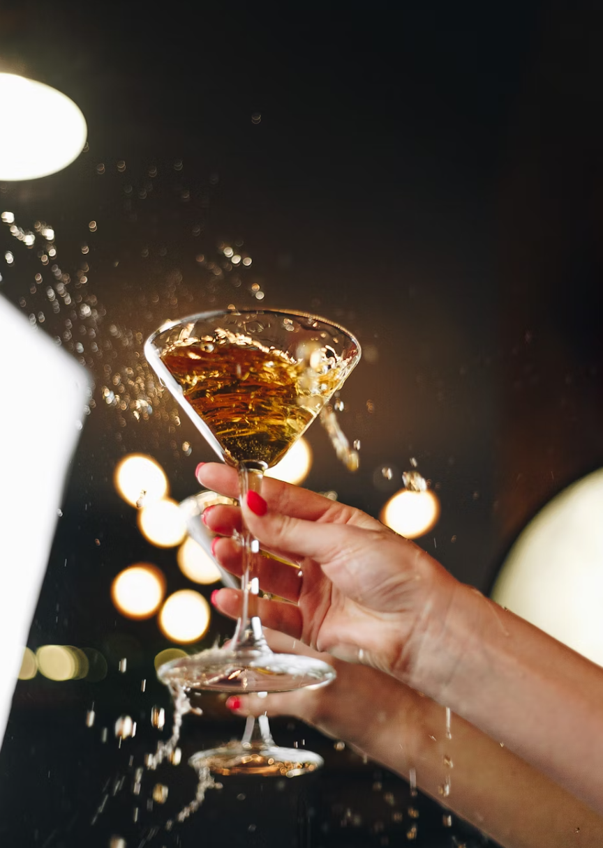 A hand with red nail polish holding a martini glass filled with a dark-colored beverage, with liquid splashing out as the glass is tilted, against a dark background with blurred lights.
