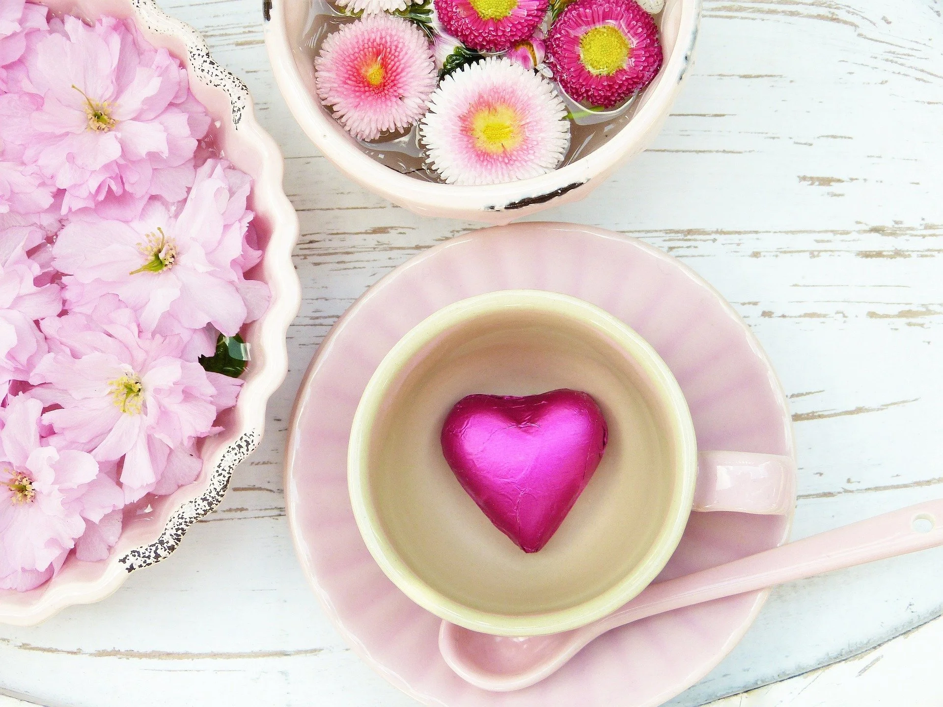 Pink and purple flowers in bowls surrounding a teacup with a pink heart-shaped object inside, on a white wooden surface.