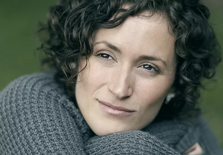 Close-up of a woman with curly hair and a gray sweater, outdoors with a blurred green background.