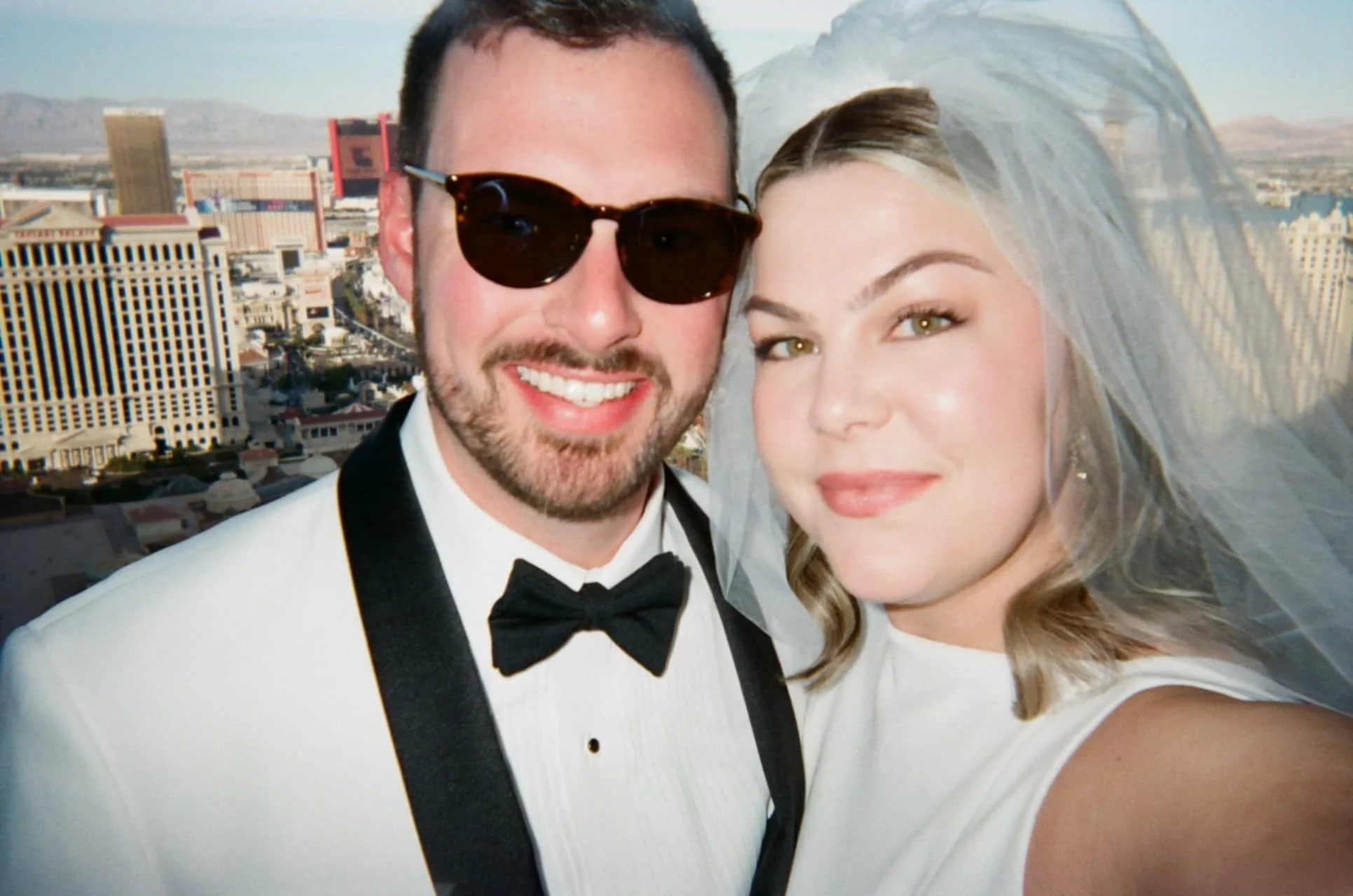 A smiling couple dressed in wedding attire taking a selfie with a cityscape background, including tall buildings and mountains.