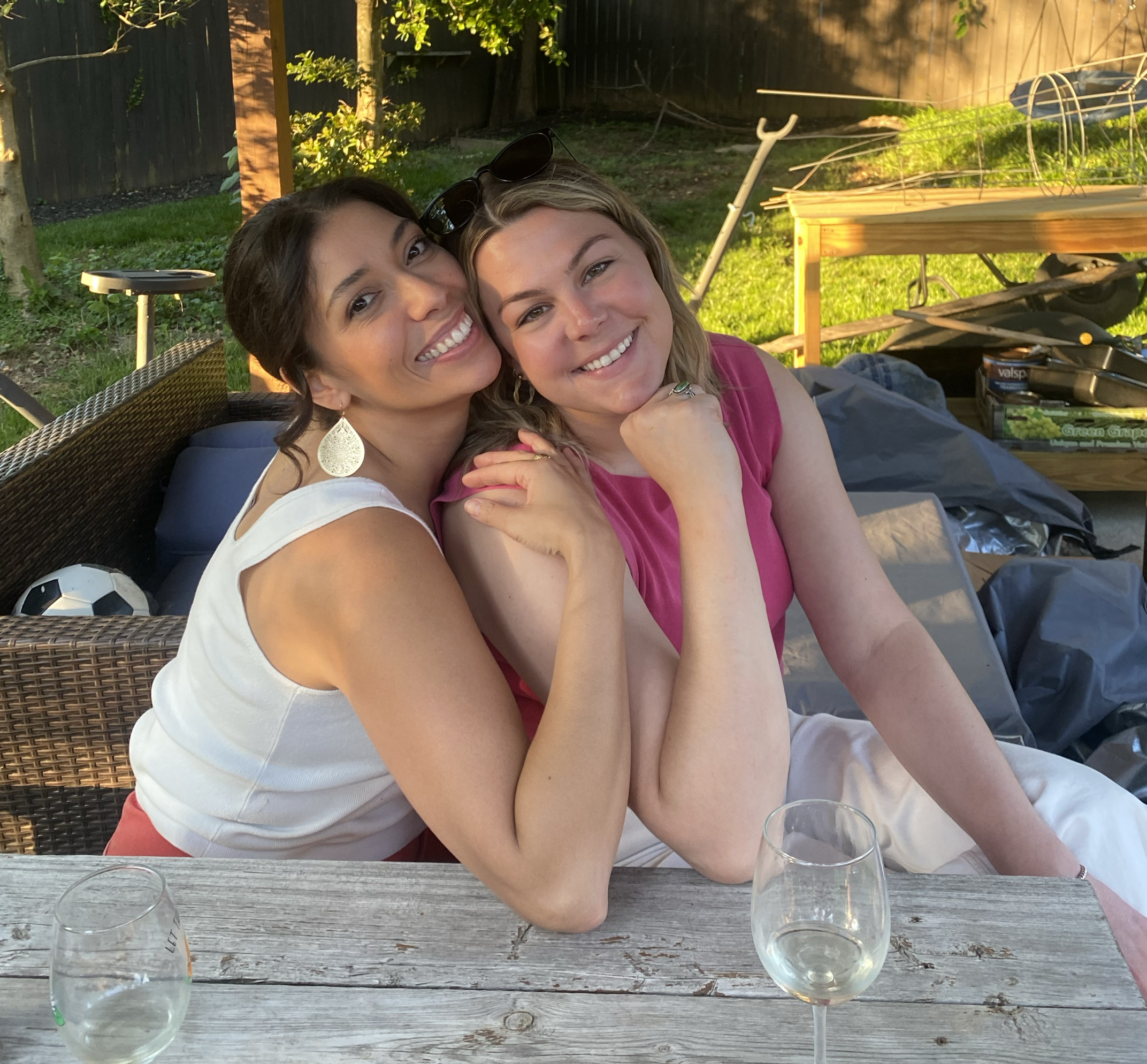 Two women smiling and resting their heads together at an outdoor gathering, with glasses of water on a wooden table in front of them and a background of a garden, fencing, and sunlight.