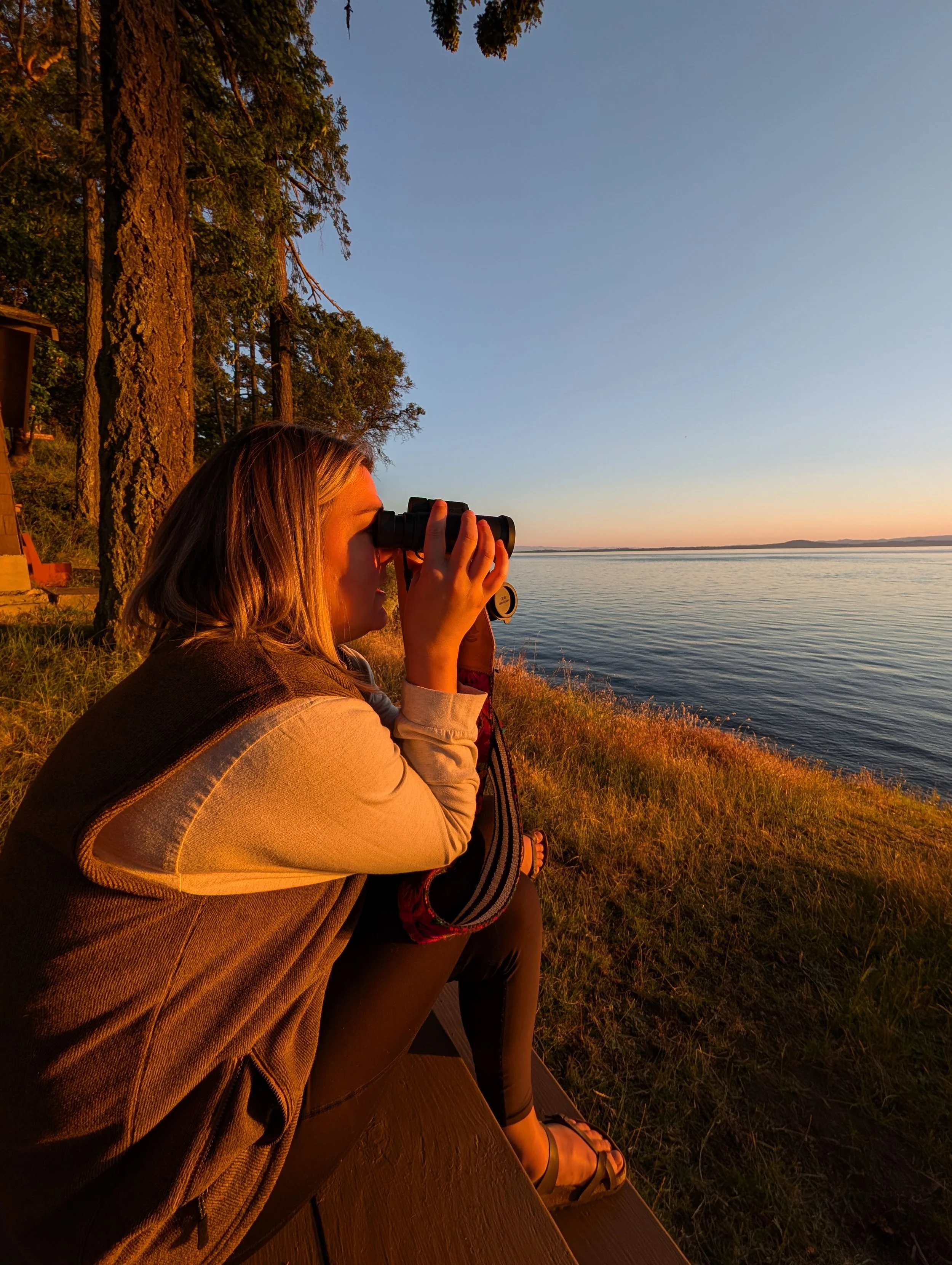 A woman sitting on a dock by a lake at sunset, gazing through binoculars with trees and a calm water body in the background.
