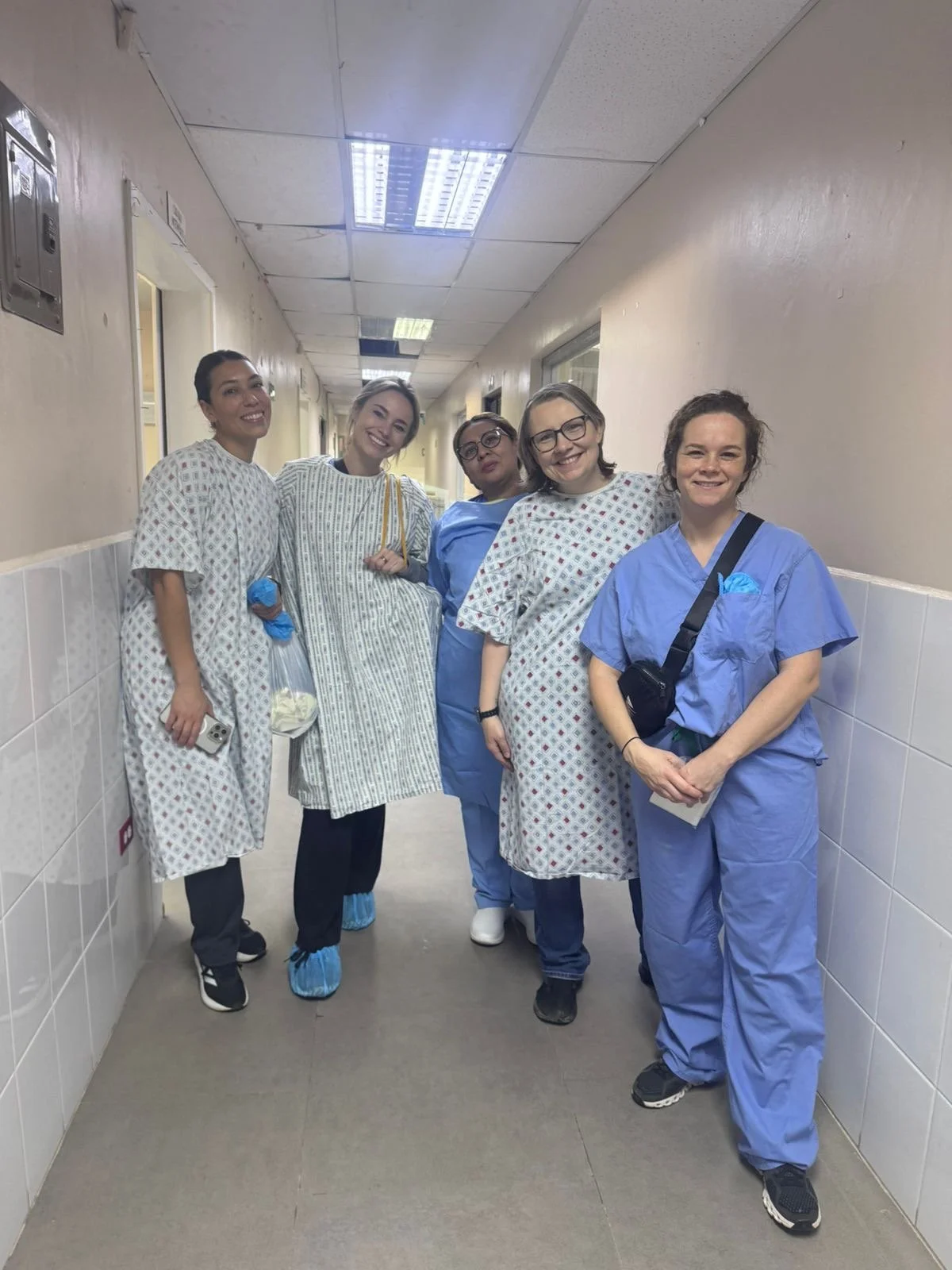 Group of five women in hospital gowns and scrubs walking down a hospital hallway, smiling at the camera.