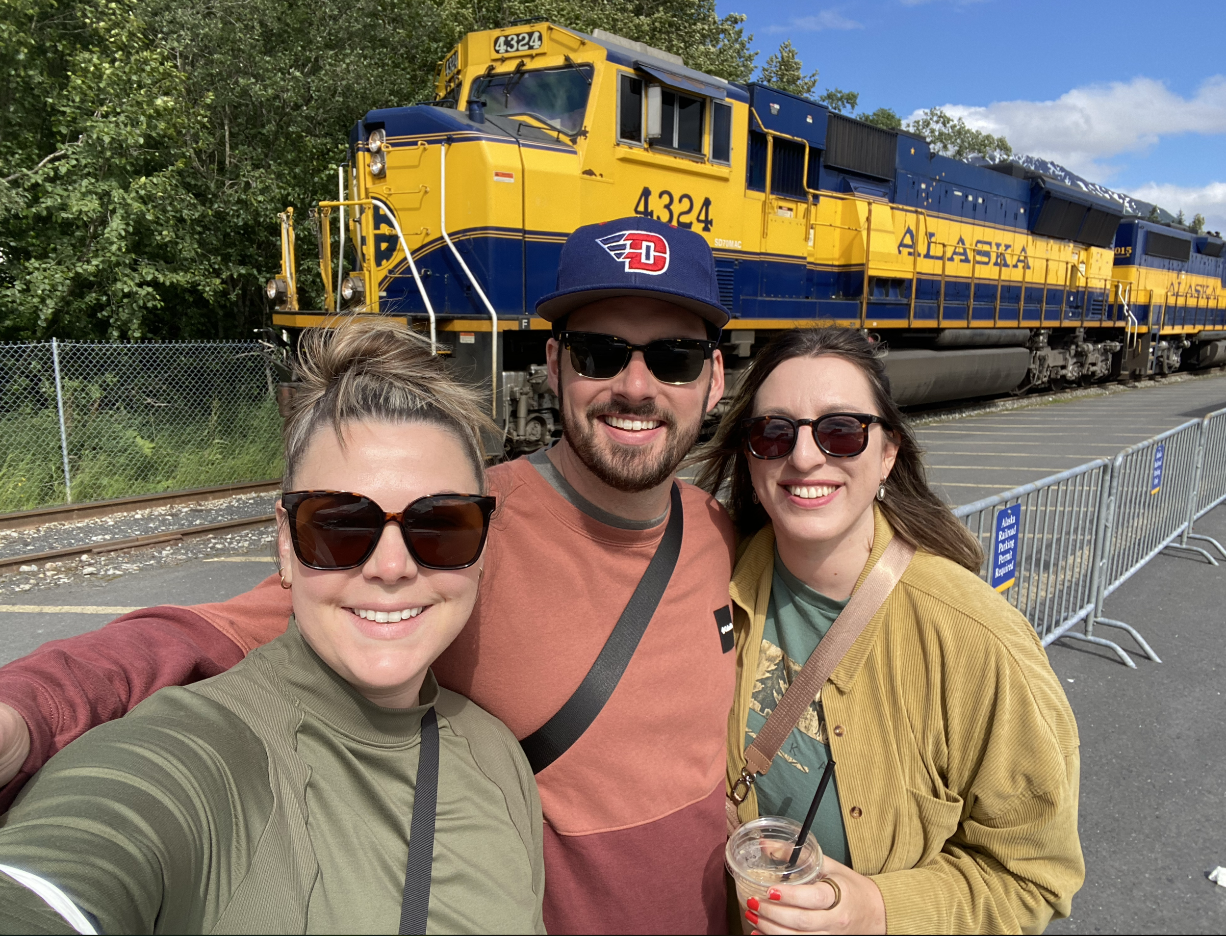 A group of four friends taking a selfie in front of a yellow and blue Alaska Railroad train, with lush green trees and blue sky in the background.