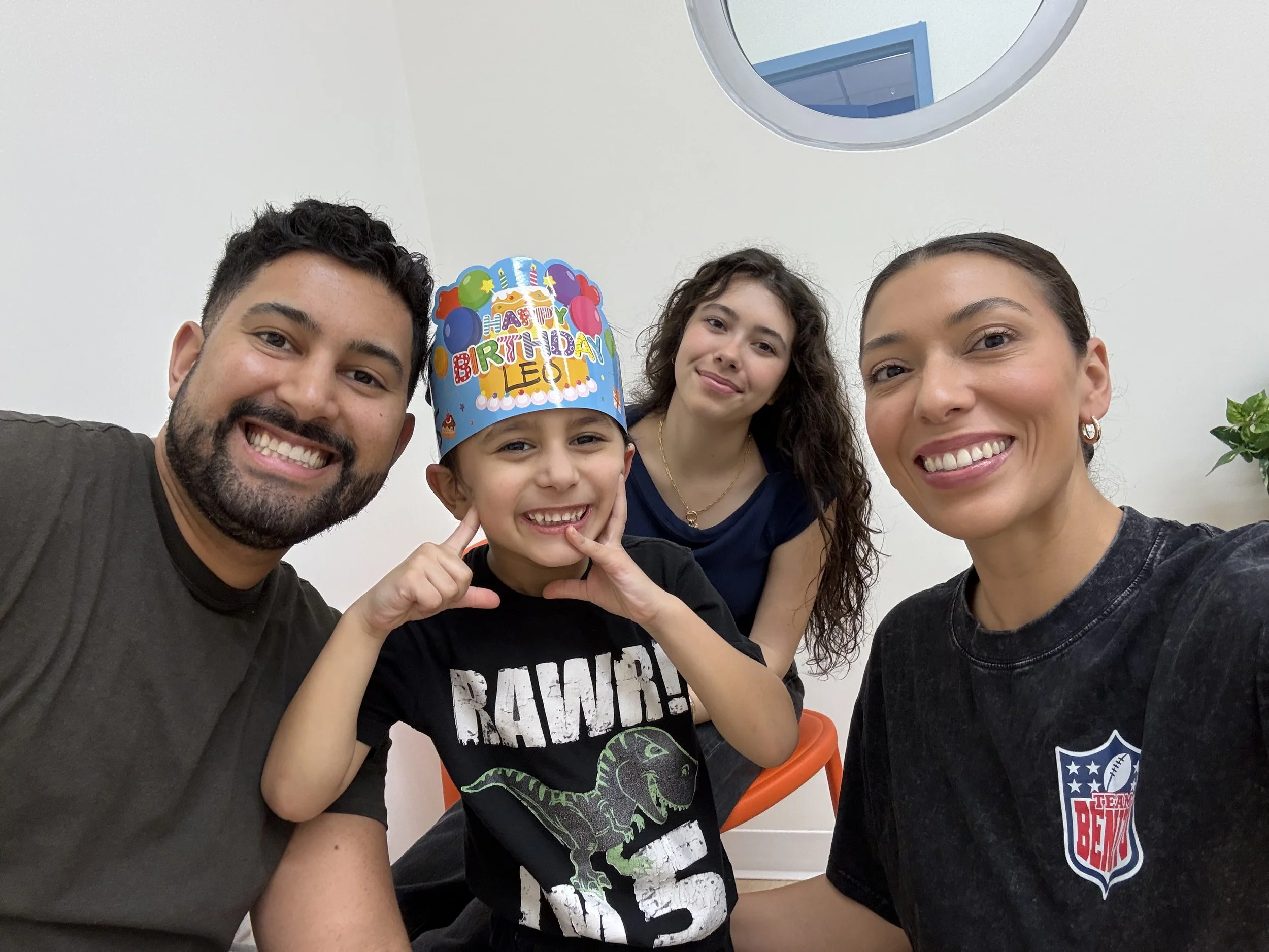 A group of four people, two women and two children, smiling and taking a selfie indoors. One child is wearing a birthday hat that says 'Happy Birthday Leo'.