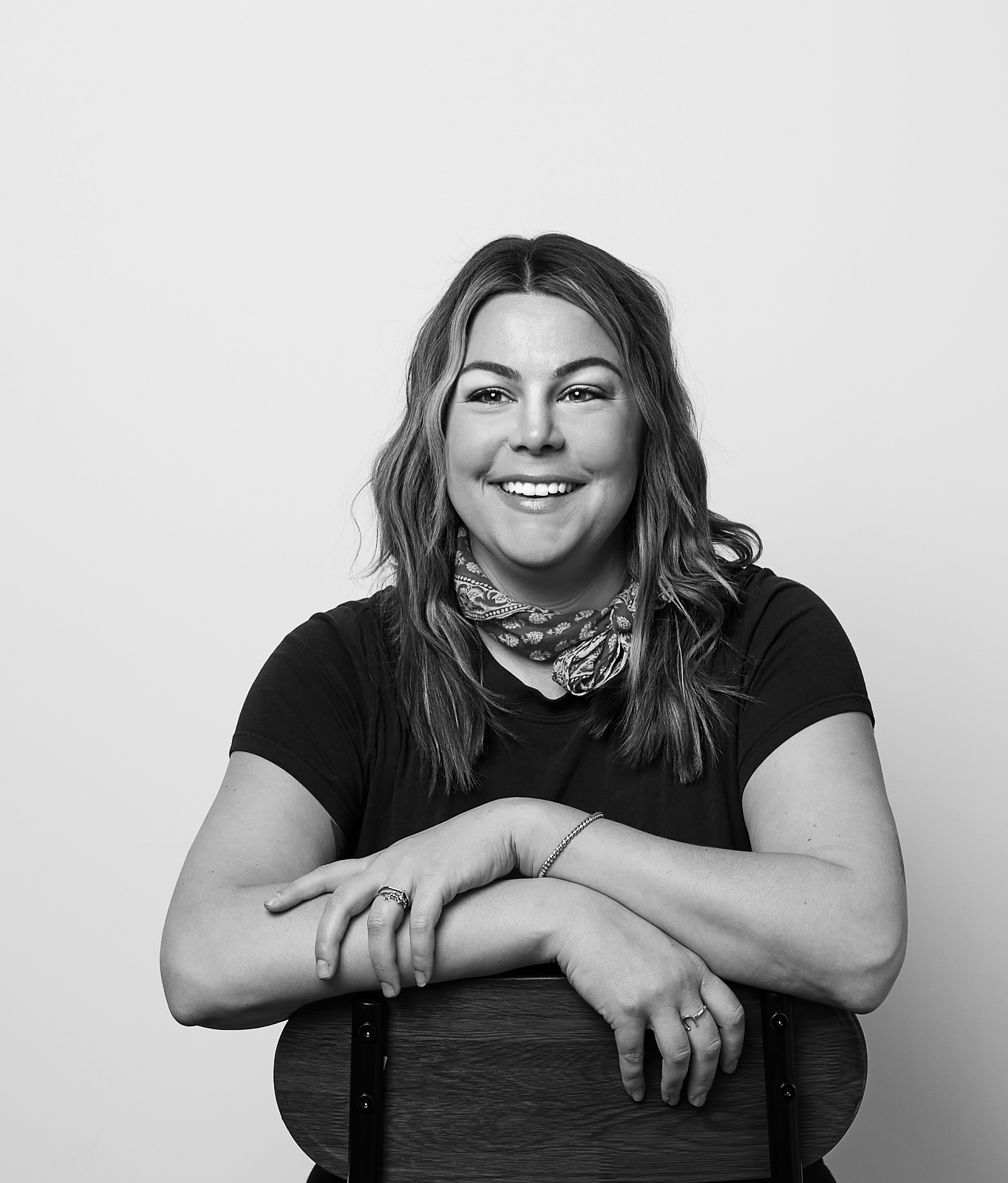 Photo of Kristen Haws | Black and white portrait of a smiling woman with wavy hair, wearing a bandana around her neck, a black t-shirt, and jewelry, sitting on a wooden chair against a plain light background.