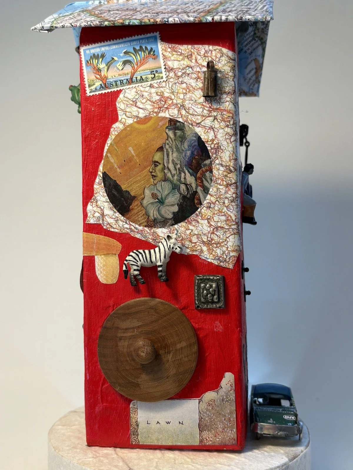 Mixed-media art piece on a red rectangular canvas featuring a postage stamp from Australia, a circular cut-out with a collage of a woman's profile, flowers, and a landscape, a small zebra figurine, a wooden disc, a metal embellishment, and a miniature toy car at the bottom, with a label that says 'LAWN'